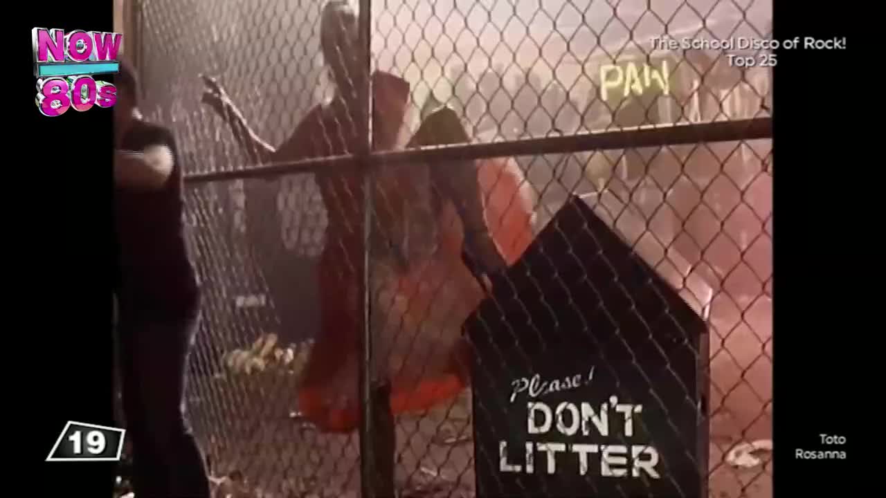 Behind the chain-link fence, a figure in a bright orange outfit is moving, arms raised. A sign warns viewers to not litter as the music video plays on Now 80s.
