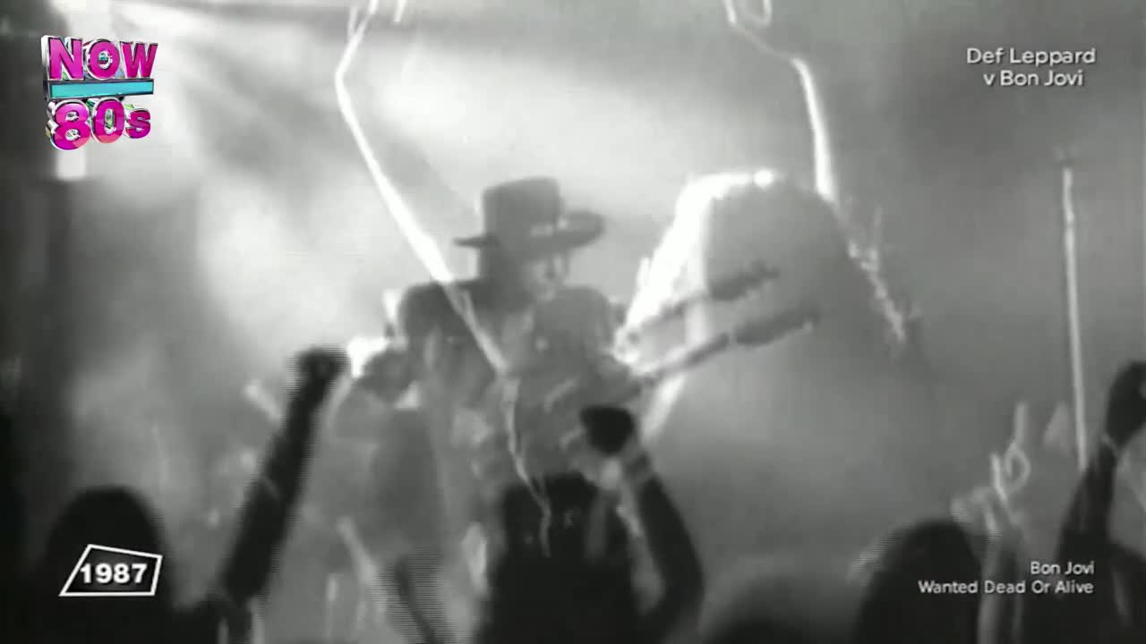 A man in a hat is silhouetted, strumming a guitar. Arms are raised in the crowd as the band plays "Wanted Dead or Alive" on Now 80s.
