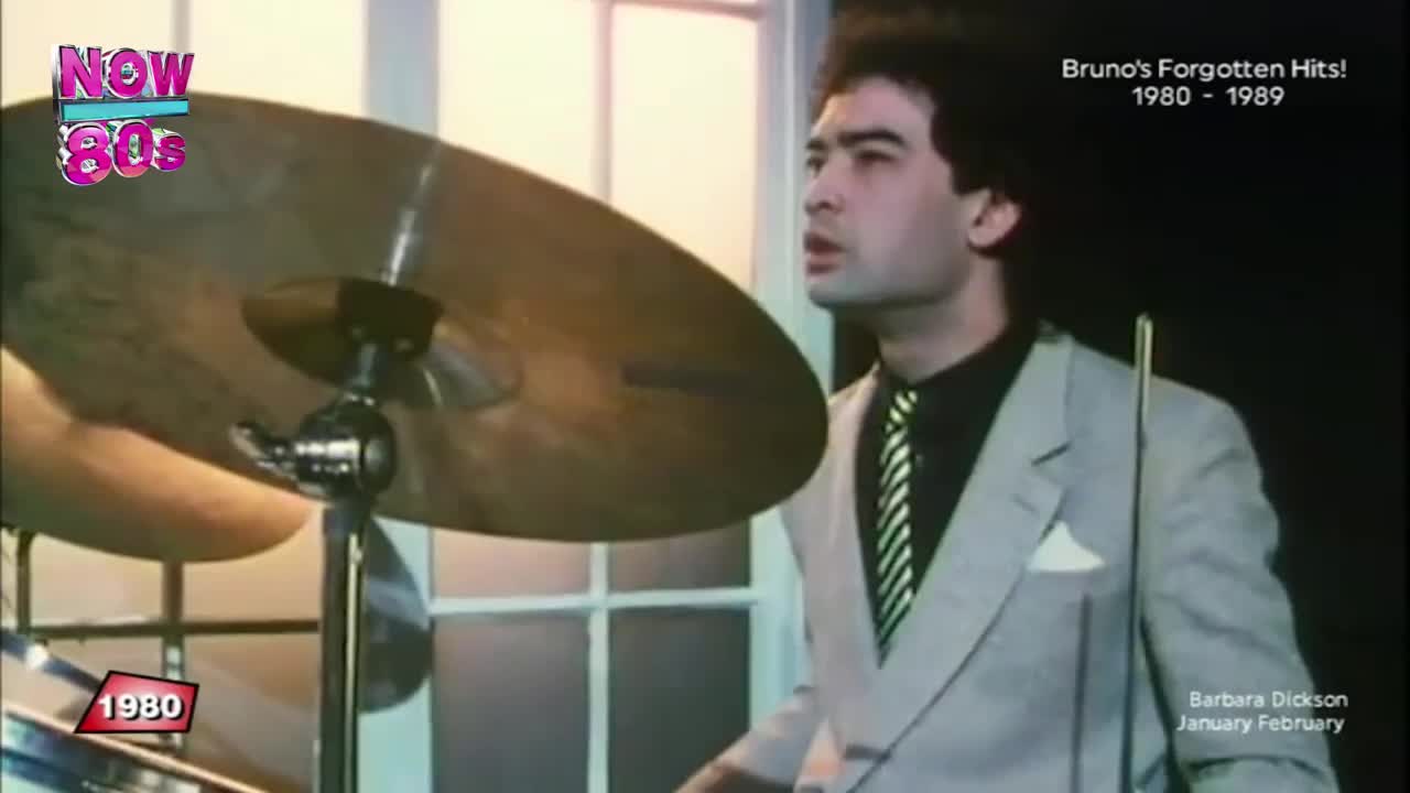 The drummer, dressed in a suit, is mid-song, his gaze fixed ahead. A cymbal gleams in the light as he prepares to strike it.
