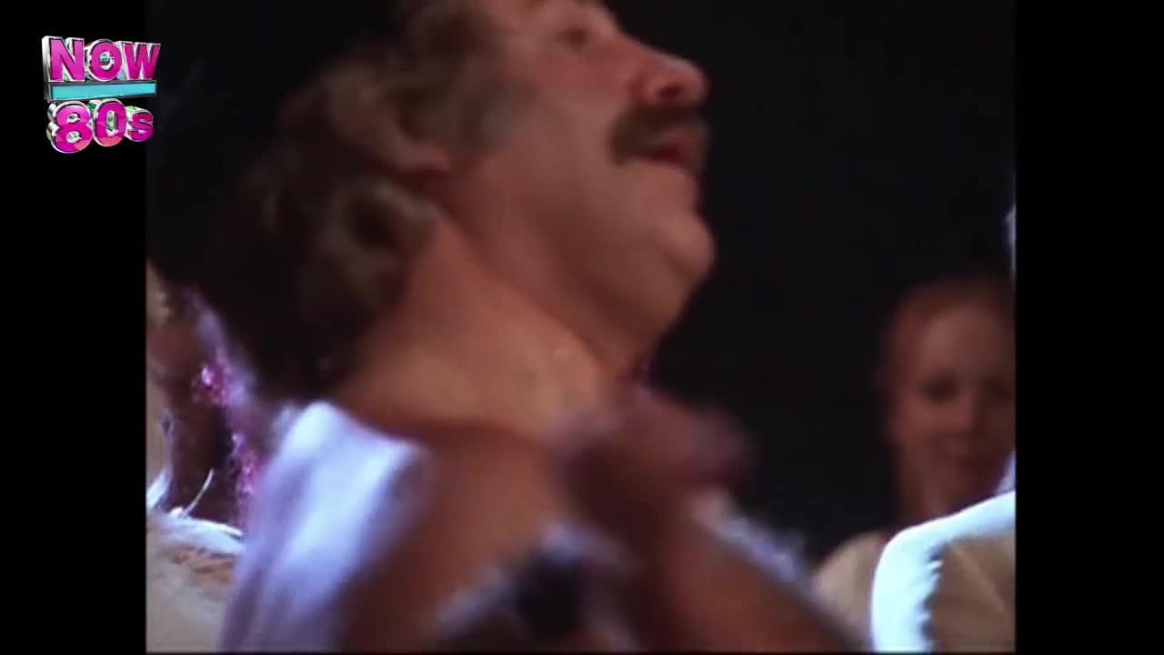 A man with a thick mustache is singing, his head thrown back. Other people are visible, but the focus is on him, illuminated by the stage lights.
