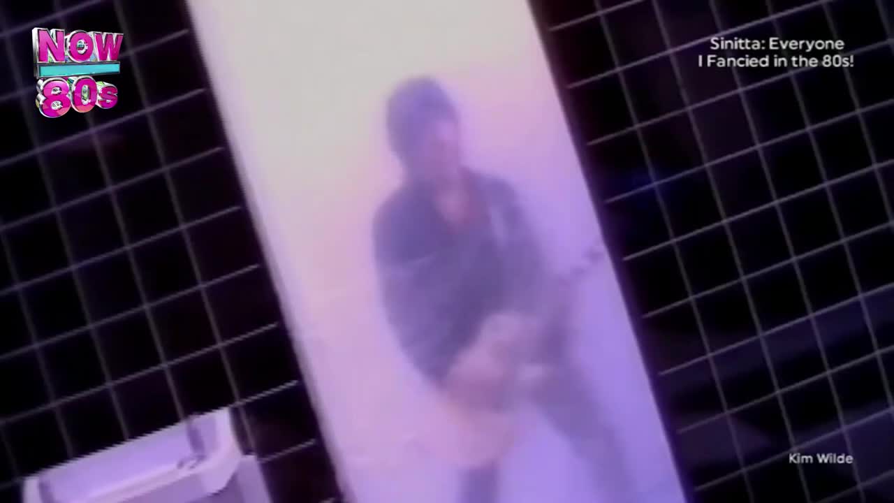 A blurry figure stands in front of a white background, likely on a music video from the 80s. The "Now 80s" logo is in the corner, and the screen is tinted with a purple hue.
