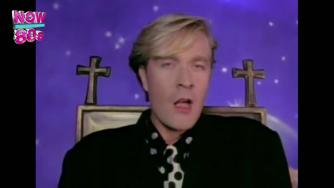 The singer, dressed in black with a polka-dot tie, is singing with a purple backdrop and crosses behind him. He's framed by a gold-colored box, and the Now 80s logo is in the corner.
