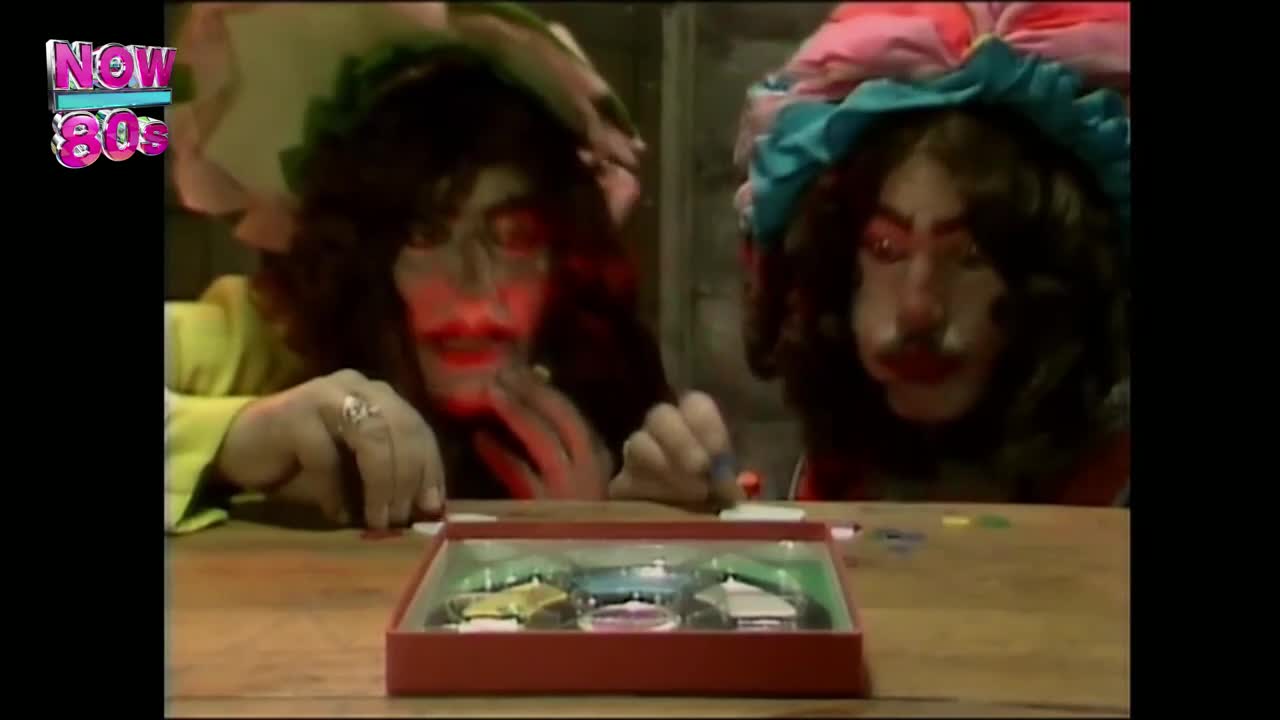 Two figures, faces heavily made up, lean over a board game. One hand reaches for a small blue piece, while the other rests on the table, adorned with a large ring.

