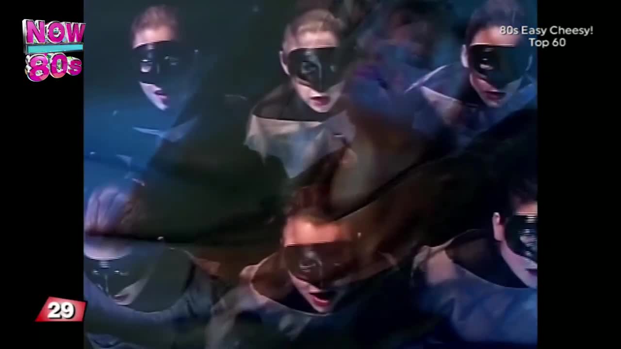 Masked figures in dark costumes are moving in a swirling pattern, their faces lit by blue light. The scene has a slightly eerie, theatrical feel, like something from a music video on Now 80s.
