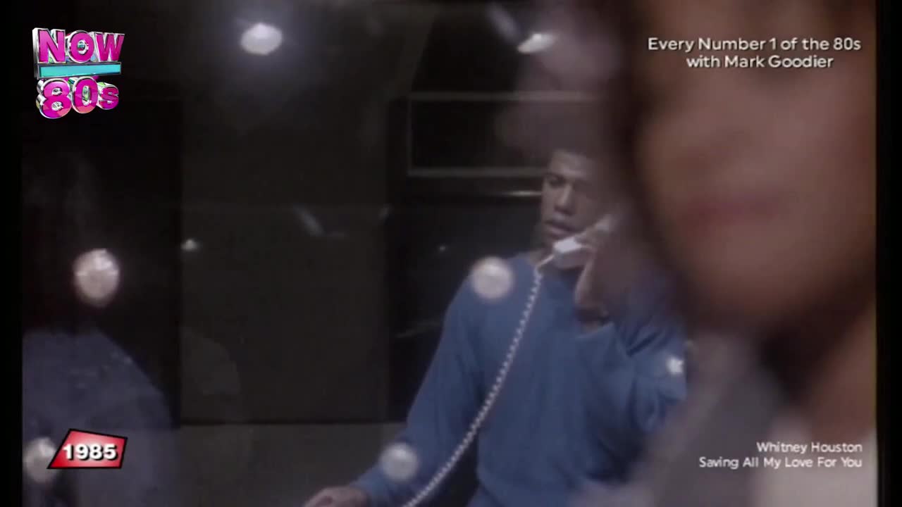 A man in a blue sweater holds a phone to his ear, his expression intent.  The Now 80s logo and the year 1985 are visible, as is the title of a Whitney Houston song.
