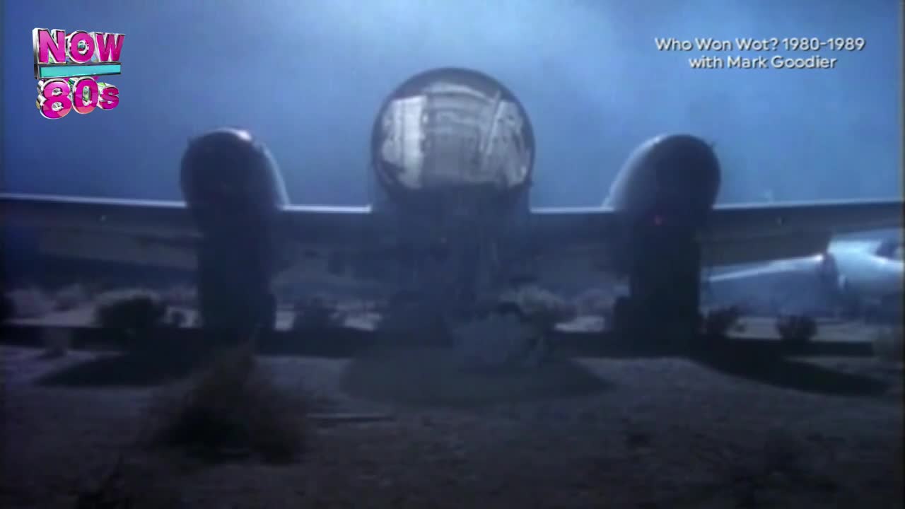 An old plane sits on the ground, its cockpit lit up against the dark sky. The scene feels like a shot from a music video on Now 80s, a throwback to the United Kingdom's past.
