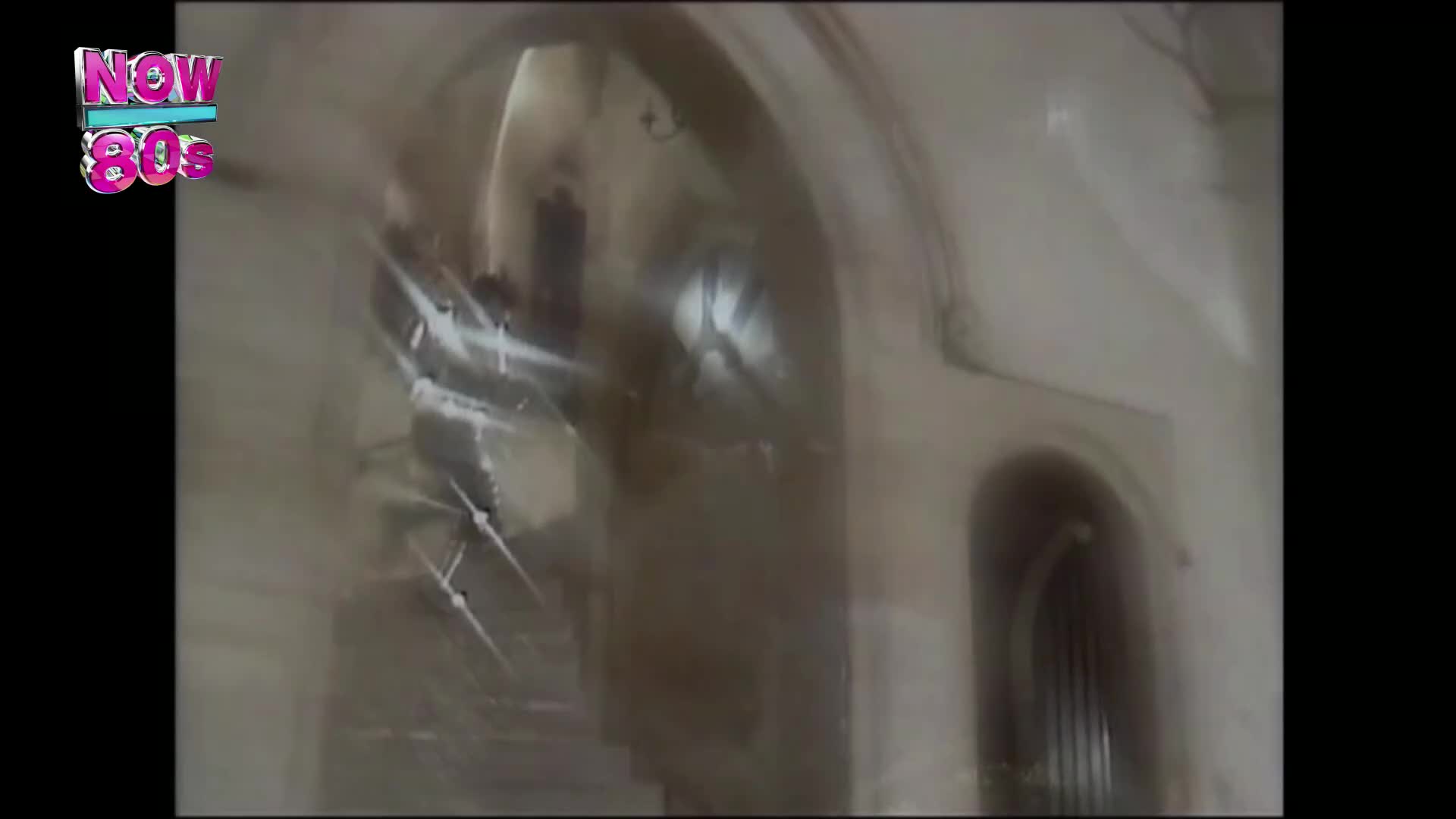 A flash of light streaks across the arched ceiling, reflecting off the stone walls. The scene, broadcast on Now 80s, is a blur of movement within a grand, old building in the United Kingdom.
