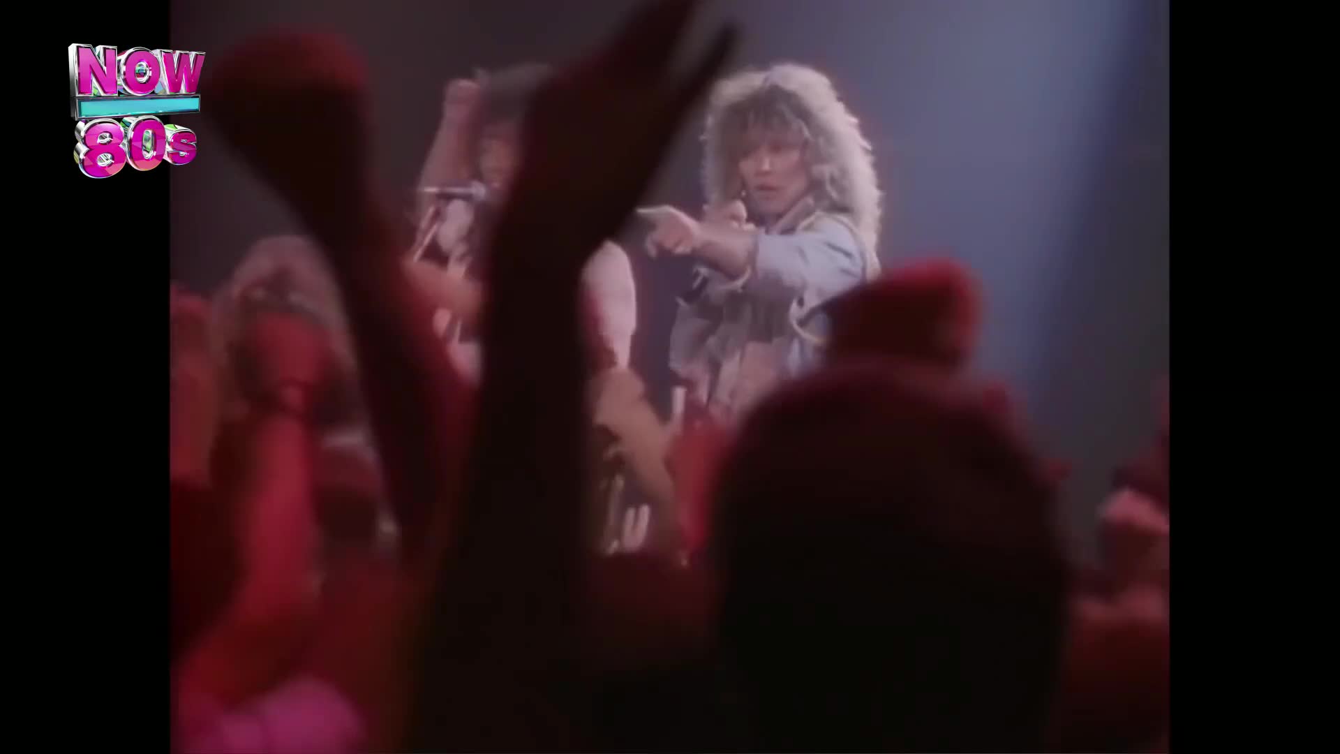 The singer with the big hair points her finger towards the crowd. Arms reach up from the audience, lit in red, as the band plays on the stage.
