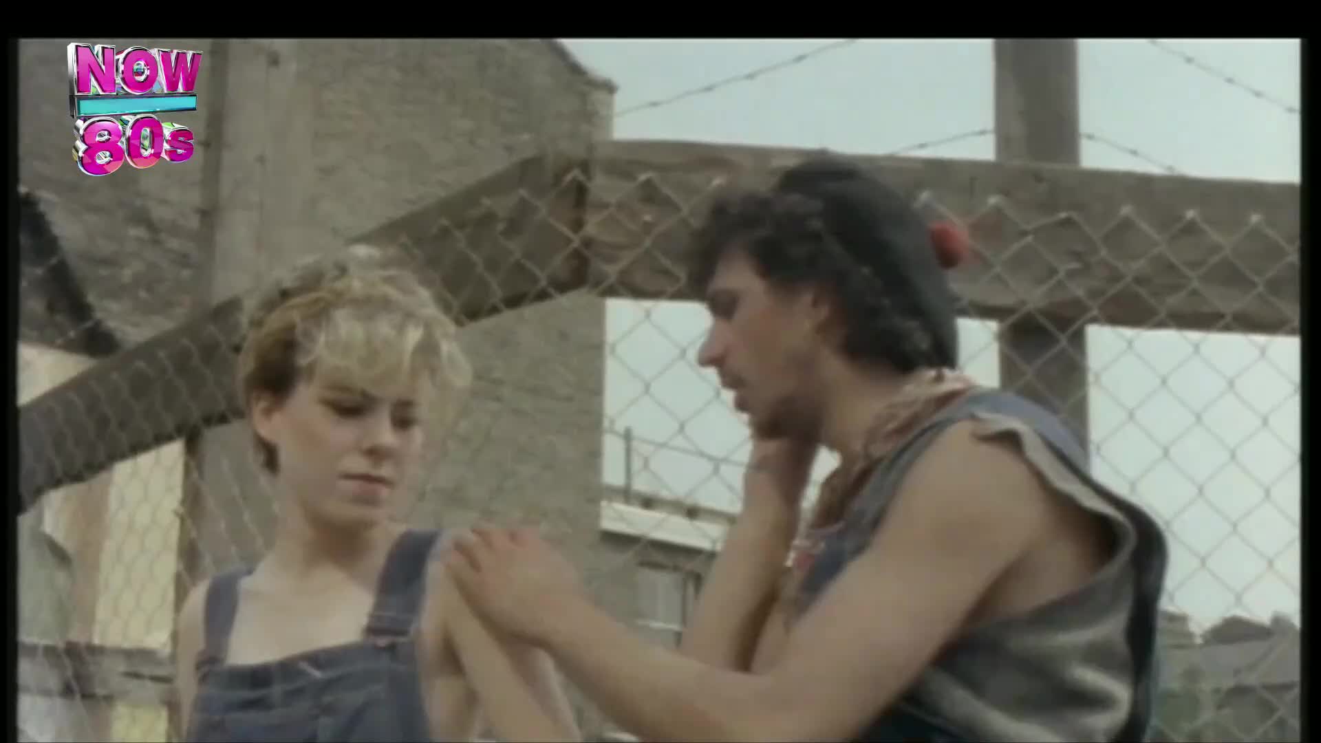 A woman in overalls looks down as a man in a sleeveless shirt touches her arm, his face turned away. Behind them, a chain-link fence and a cloudy sky set a bleak tone.
