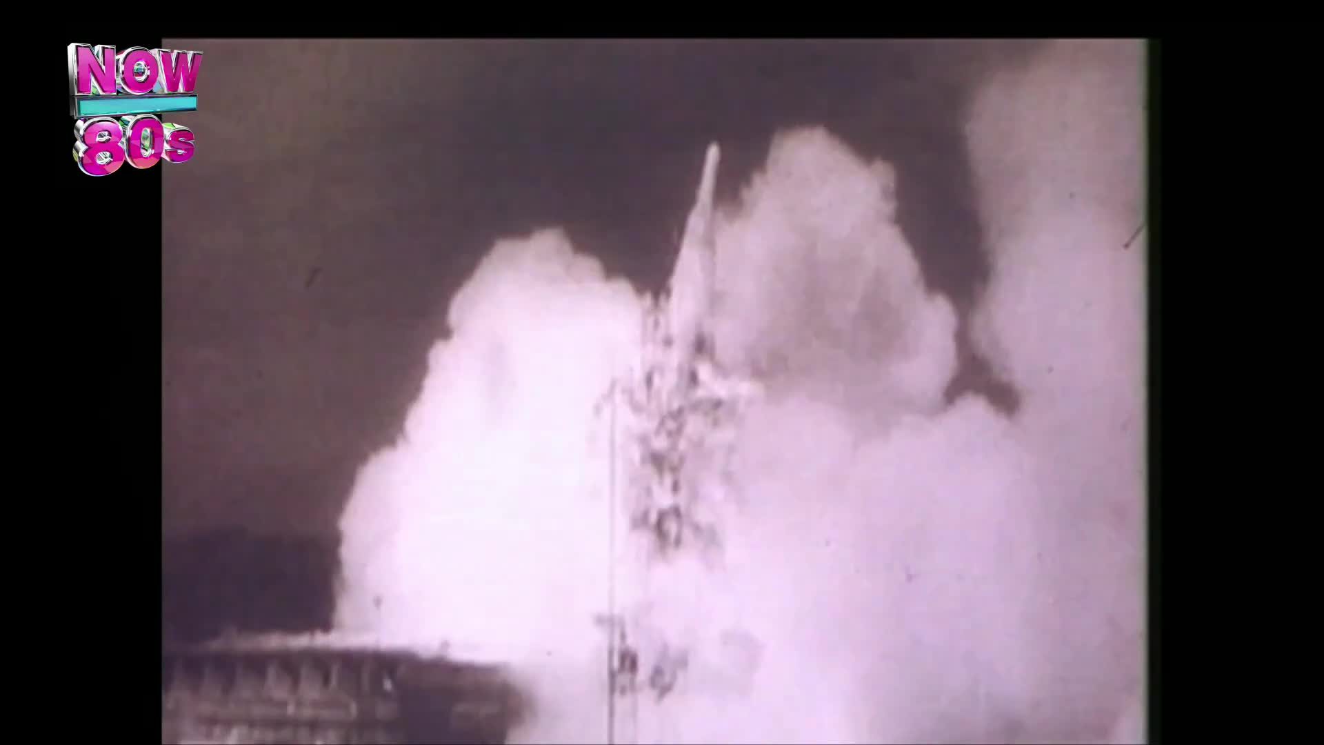 A rocket, framed by a cloud of white smoke, is ascending into the sky. The launch is captured on what looks like old film, a relic from the United Kingdom's past, perhaps from the Now 80s era.
