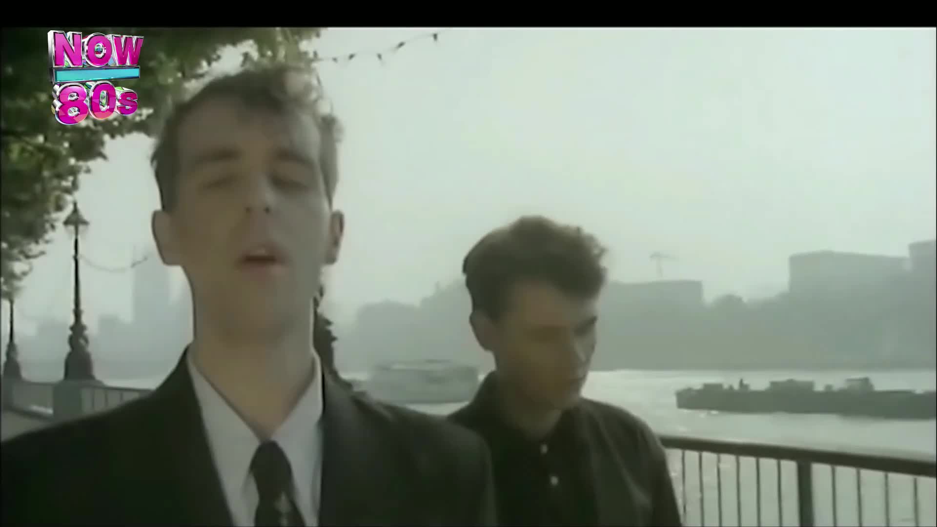 On a hazy day in the UK, two men stroll along a riverbank. One in a suit sings, while the other walks behind him, looking at the water.
