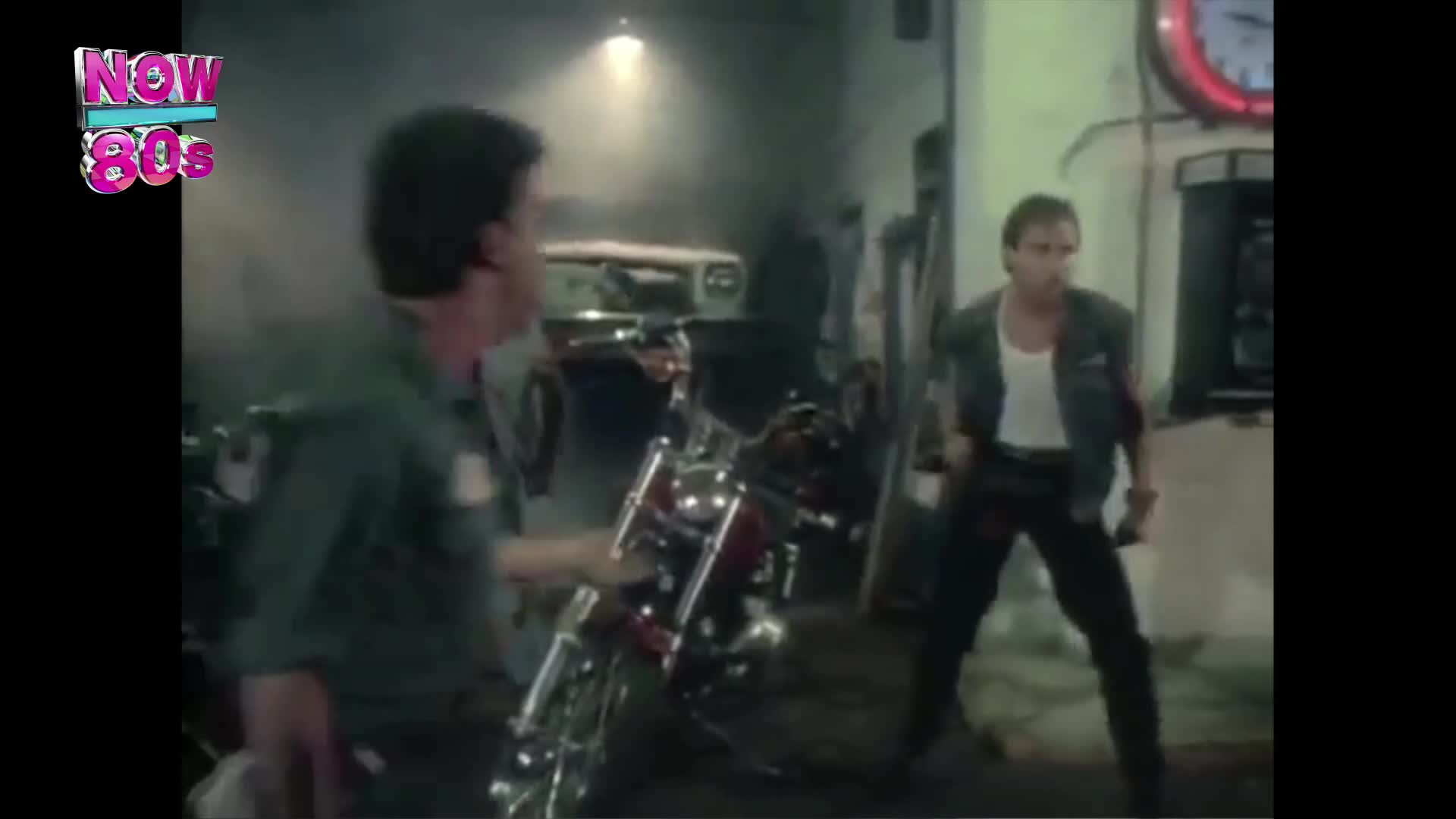 A man in a green shirt gestures towards a red motorcycle, while another man in a denim jacket stands ready, arms at his sides. The scene unfolds in a dimly lit garage, likely from a music video on Now 80s.
