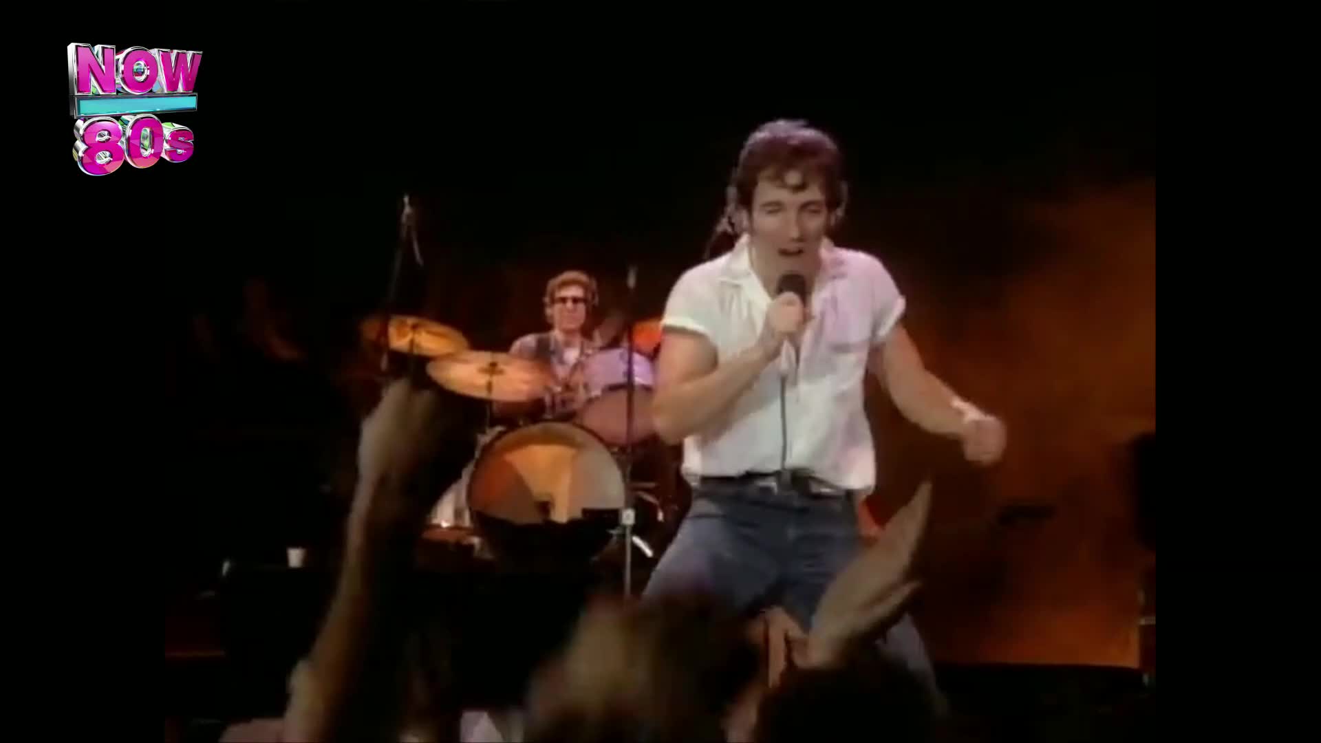 The singer, in a white shirt and jeans, is belting into a microphone, leaning into the crowd. Behind him, the drummer hits the cymbals, keeping the beat.
