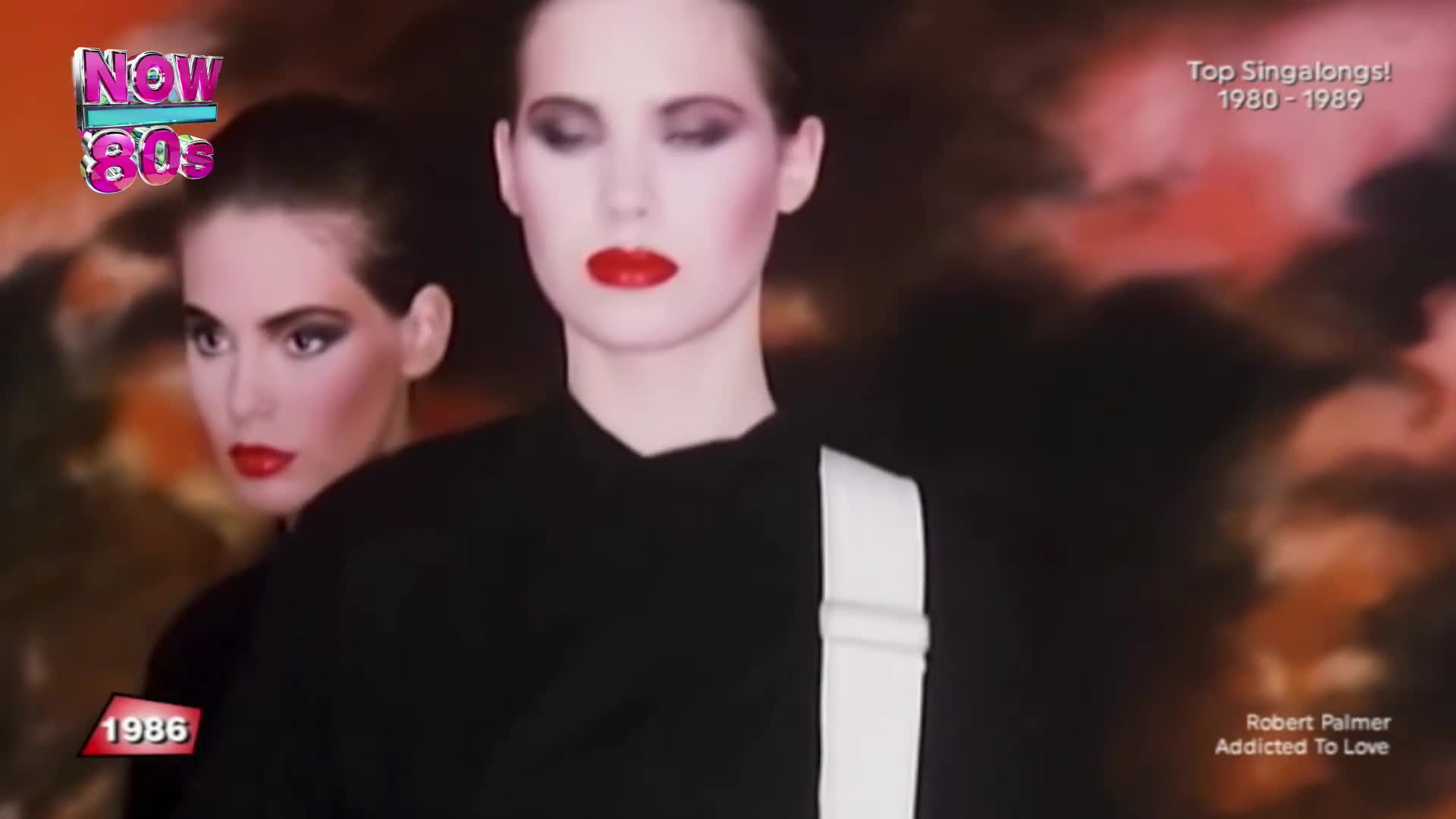 Two women with severe makeup and bright red lipstick stand in front of a hazy backdrop. One wears a black top with a white strap, while the other is partially obscured behind her.
