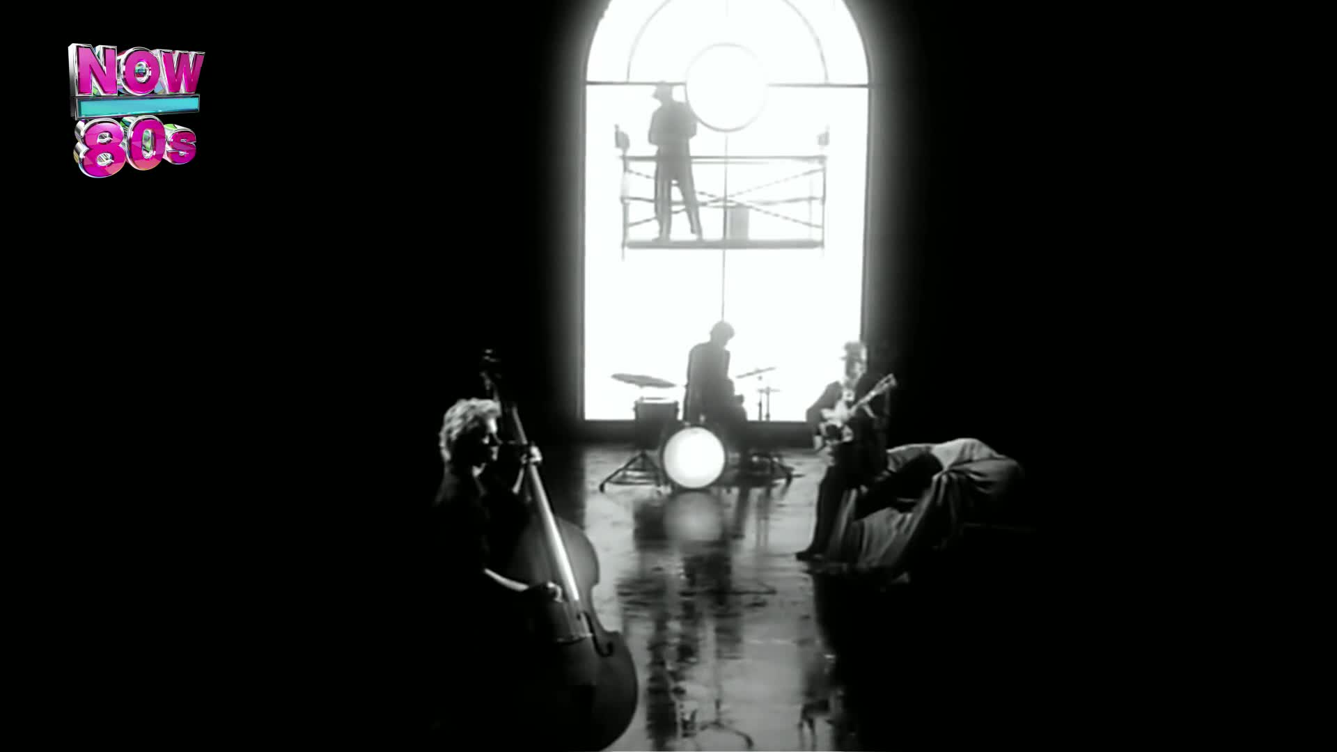A band plays in a stark, dark space, with a drummer and guitarist seated, and a bassist standing. Above them, a figure on scaffolding appears to be playing a large instrument.
