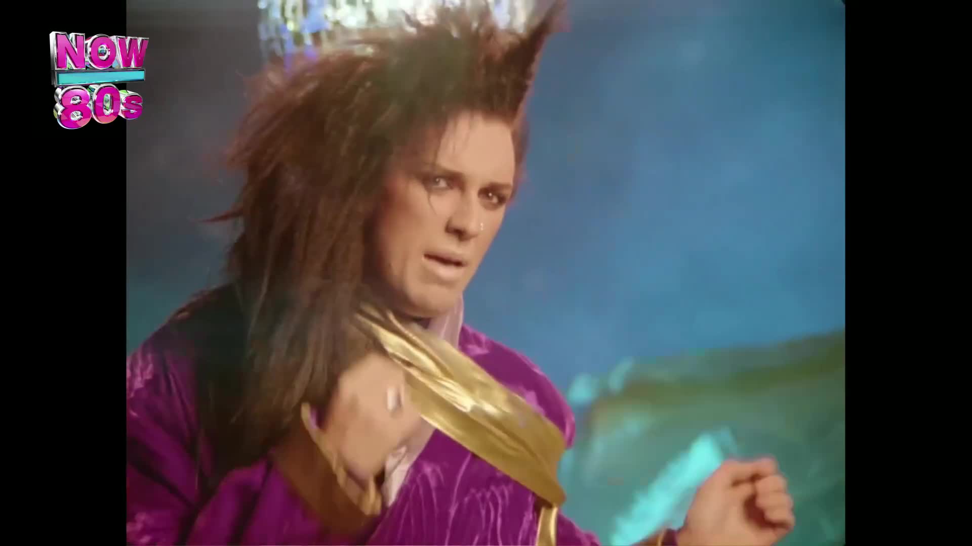 The singer, with dramatic hair, gestures with one hand while looking off to the side. He's wearing a purple outfit with a gold collar, and a glittering disco ball hangs in the background.
