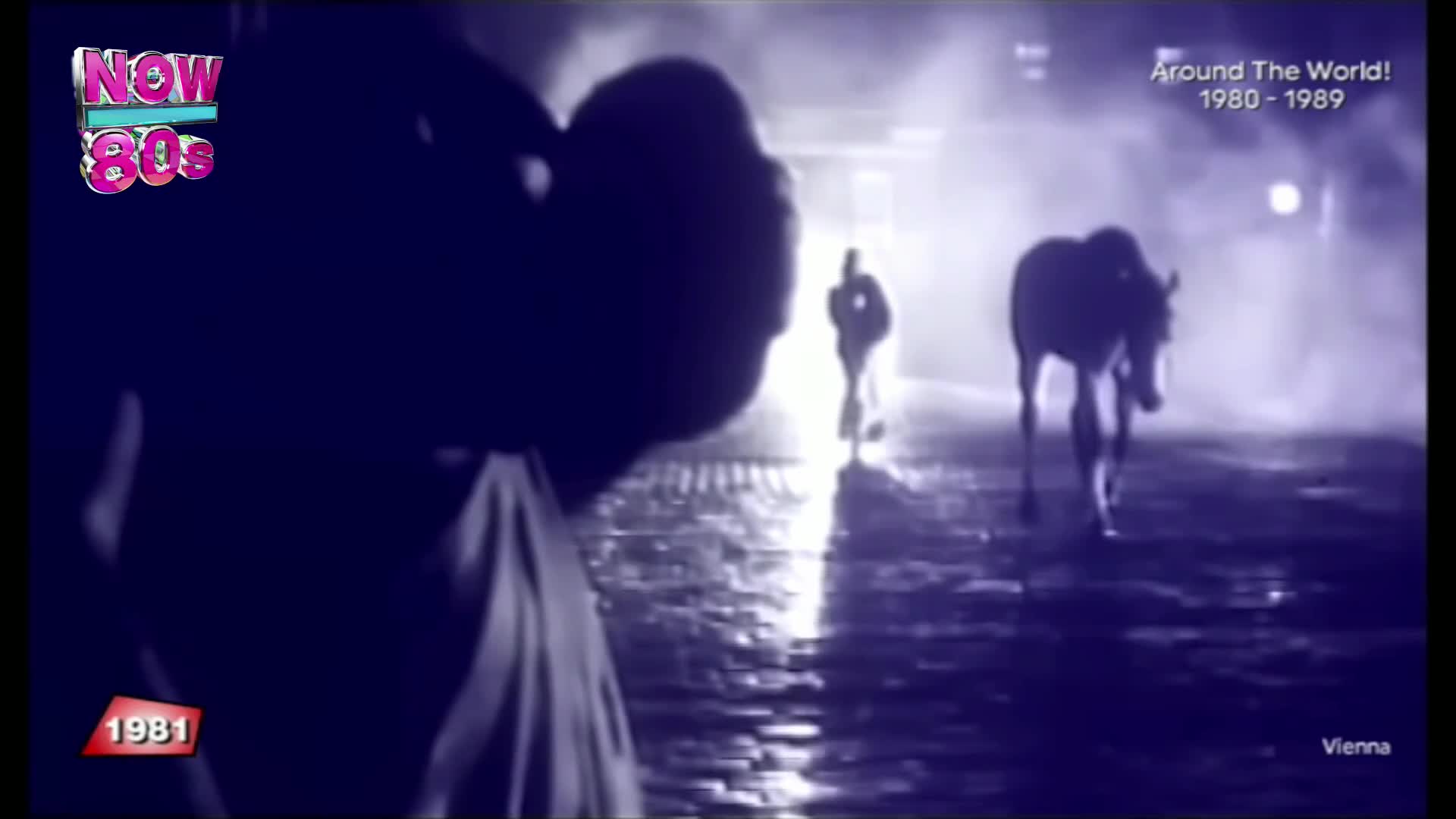 A horse walks slowly towards the camera on a wet street, with a person further down the road. The scene is bathed in a cool, misty light.
