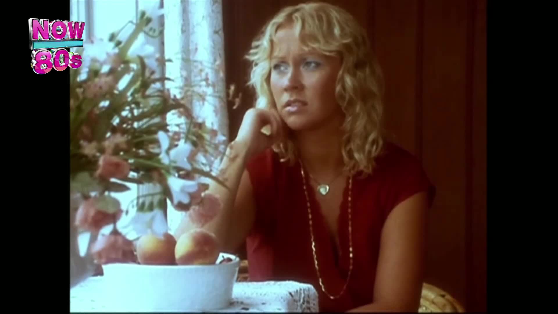 A woman with blonde hair, wearing a red top and a gold necklace, sits at a table, looking thoughtful. Peaches rest in a white bowl, and a floral arrangement sits behind her.
