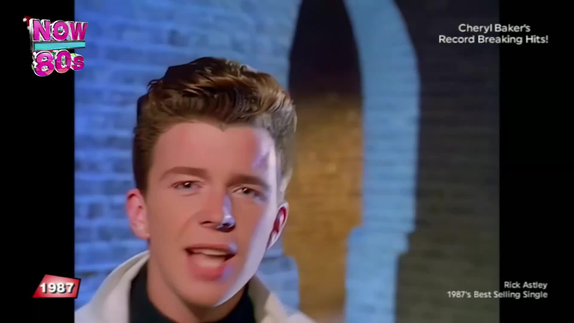 Rick Astley is singing, his mouth open as he looks straight at the camera. The background is a brick wall lit with blue and yellow light.
