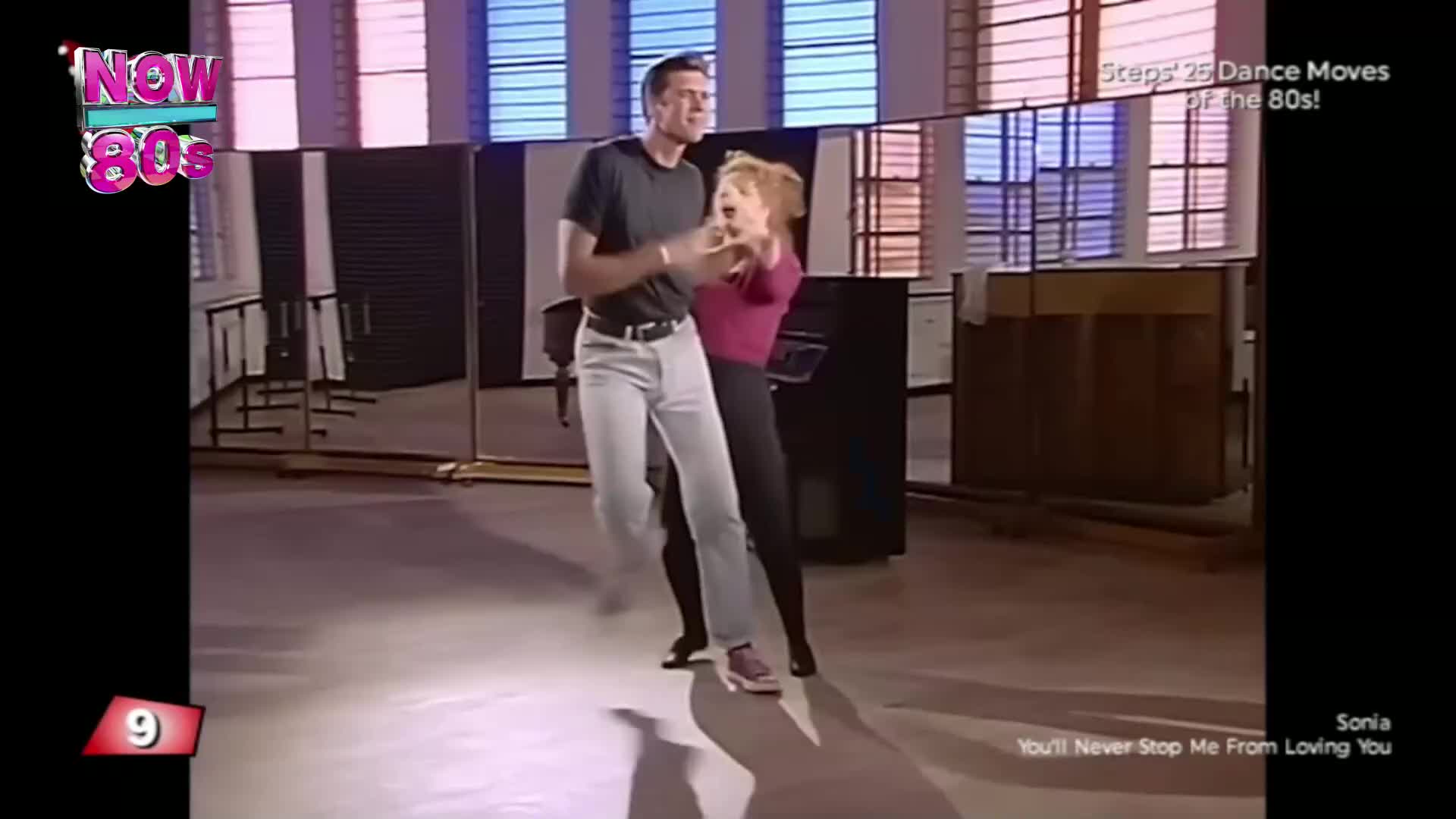A man in a black t-shirt and light jeans leads a woman in a pink top, both mid-step. They're in a studio, probably in the UK, with the Now 80s logo in the corner, and it looks like they're demonstrating a dance move.
