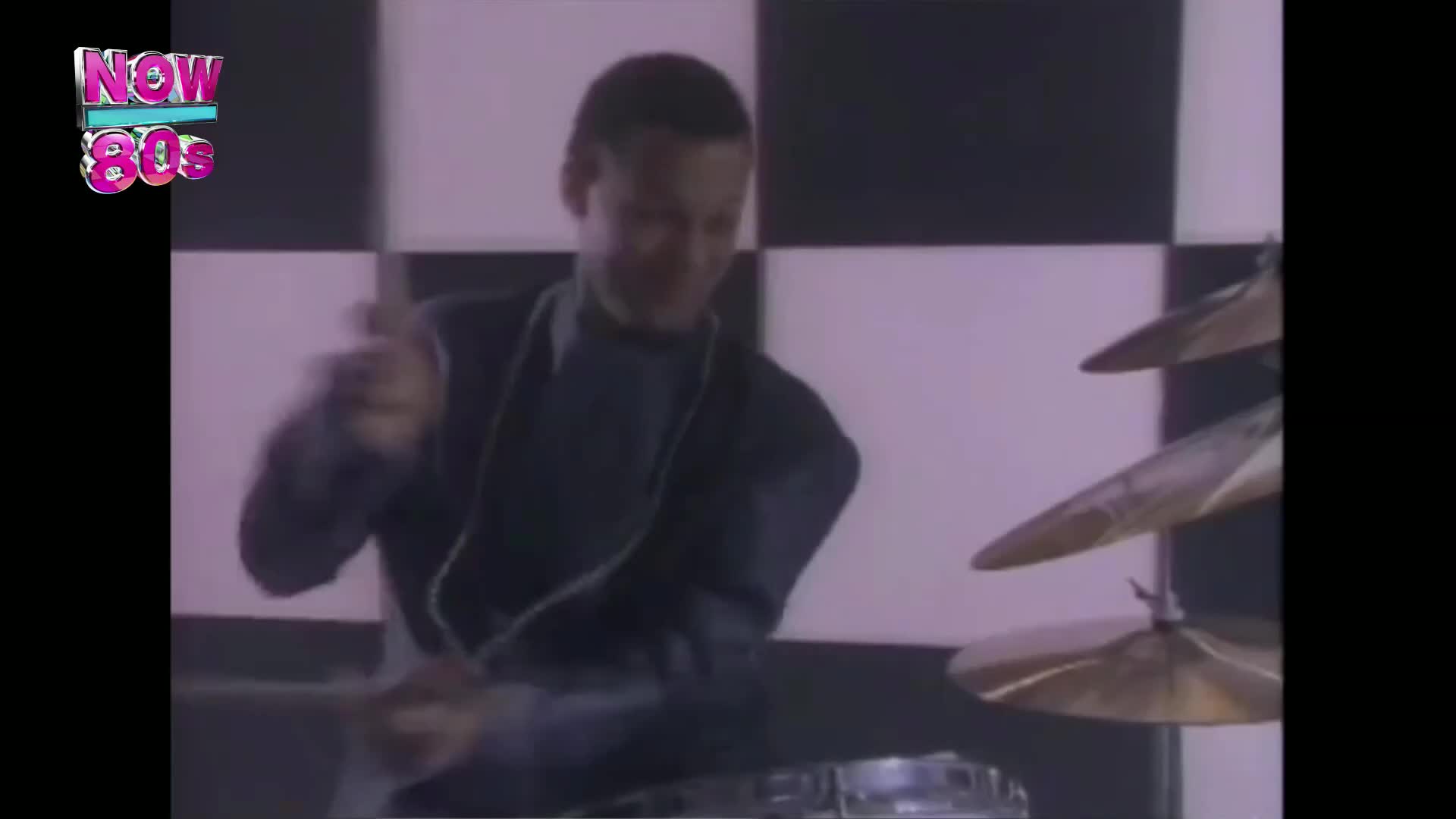 The drummer's arms are a blur as he strikes the cymbals, the sticks a flash of white against his dark suit. Behind him, a checkerboard backdrop adds a stark contrast to the scene.
