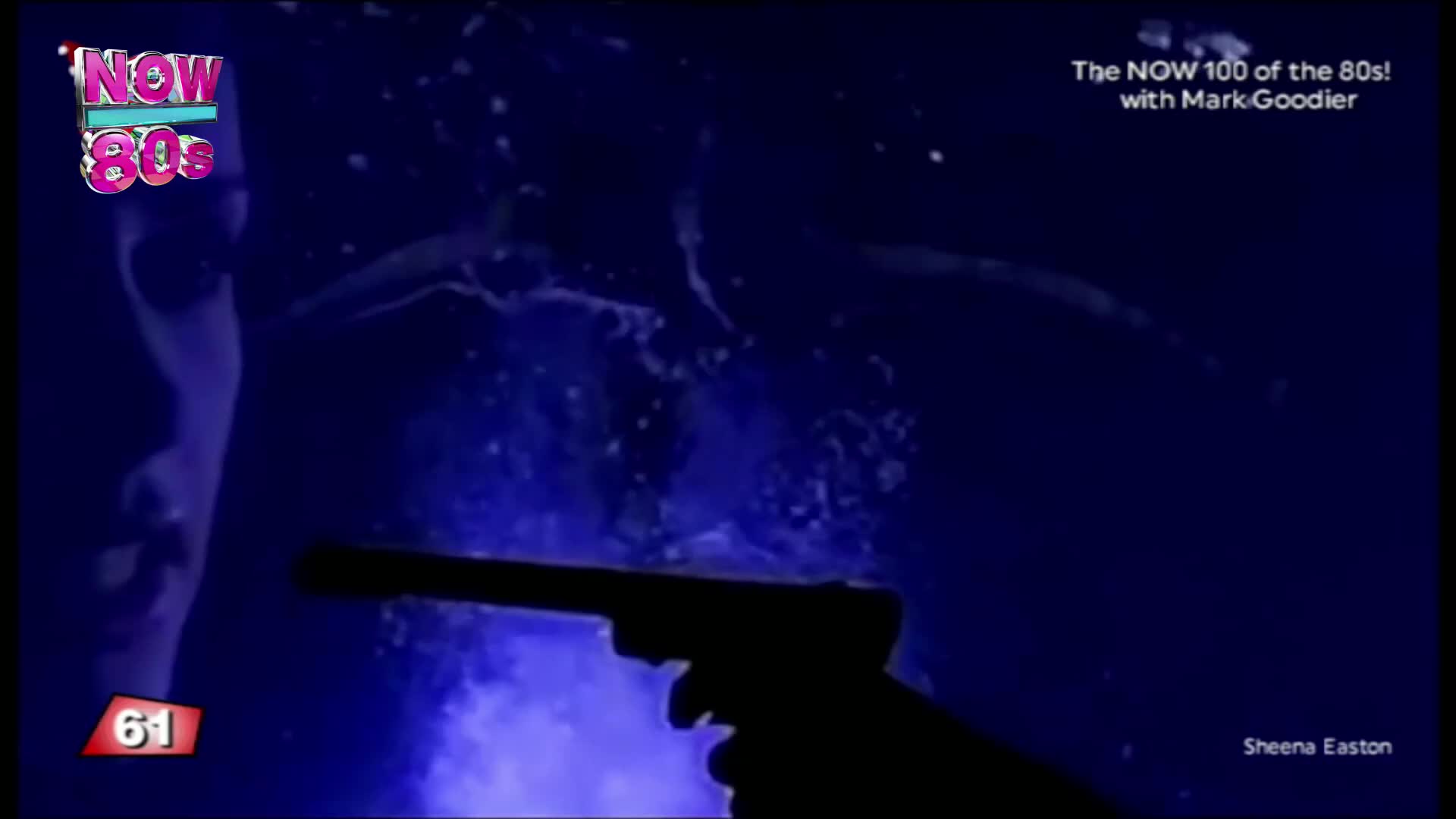 A silhouetted hand aims a gun towards a figure underwater. Water splashes around the figure, as the Now 80s logo appears in the corner.
