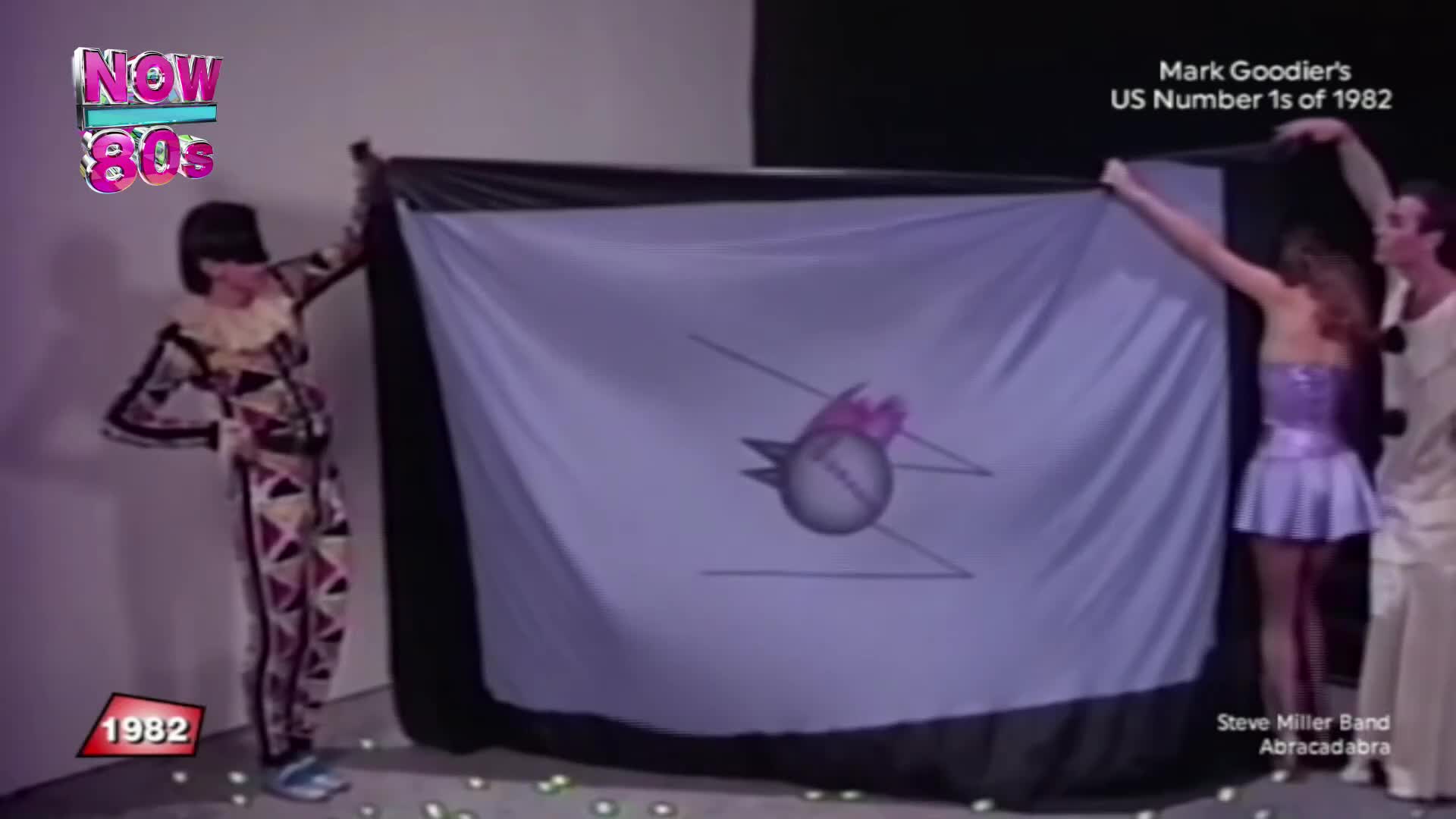 A person in a patterned jumpsuit stands to the left, while two others on the right hold up a large flag with a logo on it. The flag is being displayed on a set for a show on Now 80s.
