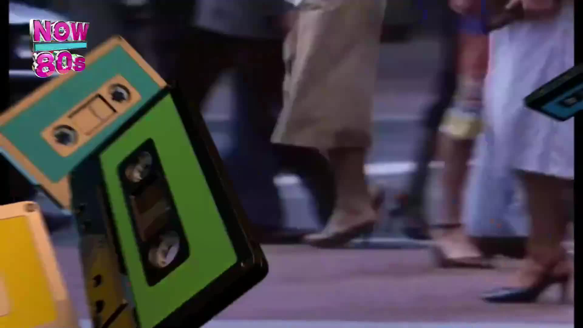 Cassette tapes in bright colours float in front of a busy street scene. People in heels and skirts walk past, their legs the only visible part of their bodies.
