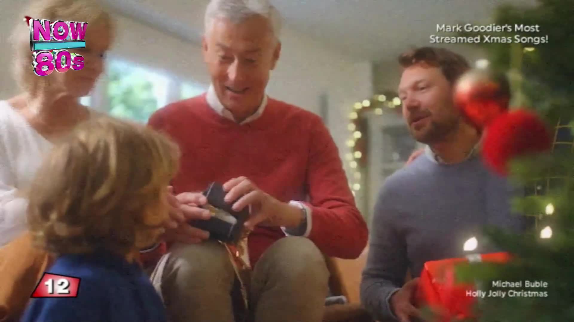 A man in a red sweater carefully opens a small, dark box. A young boy and two other adults watch intently, gathered around a Christmas tree.
