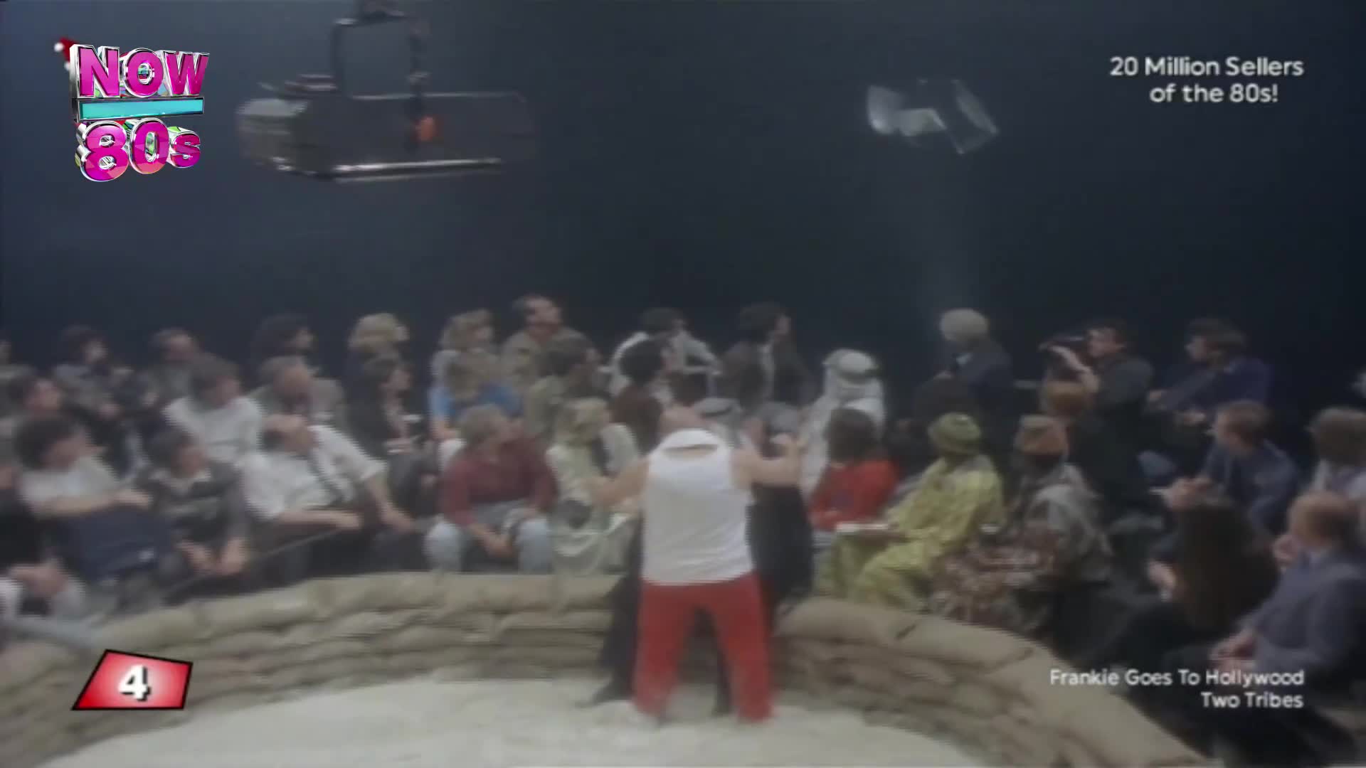A man in a white vest and red trousers stands in a sand-filled pit, arms raised, facing the audience. Above, a camera hangs suspended, and the "Now 80s" logo is visible in the corner.
