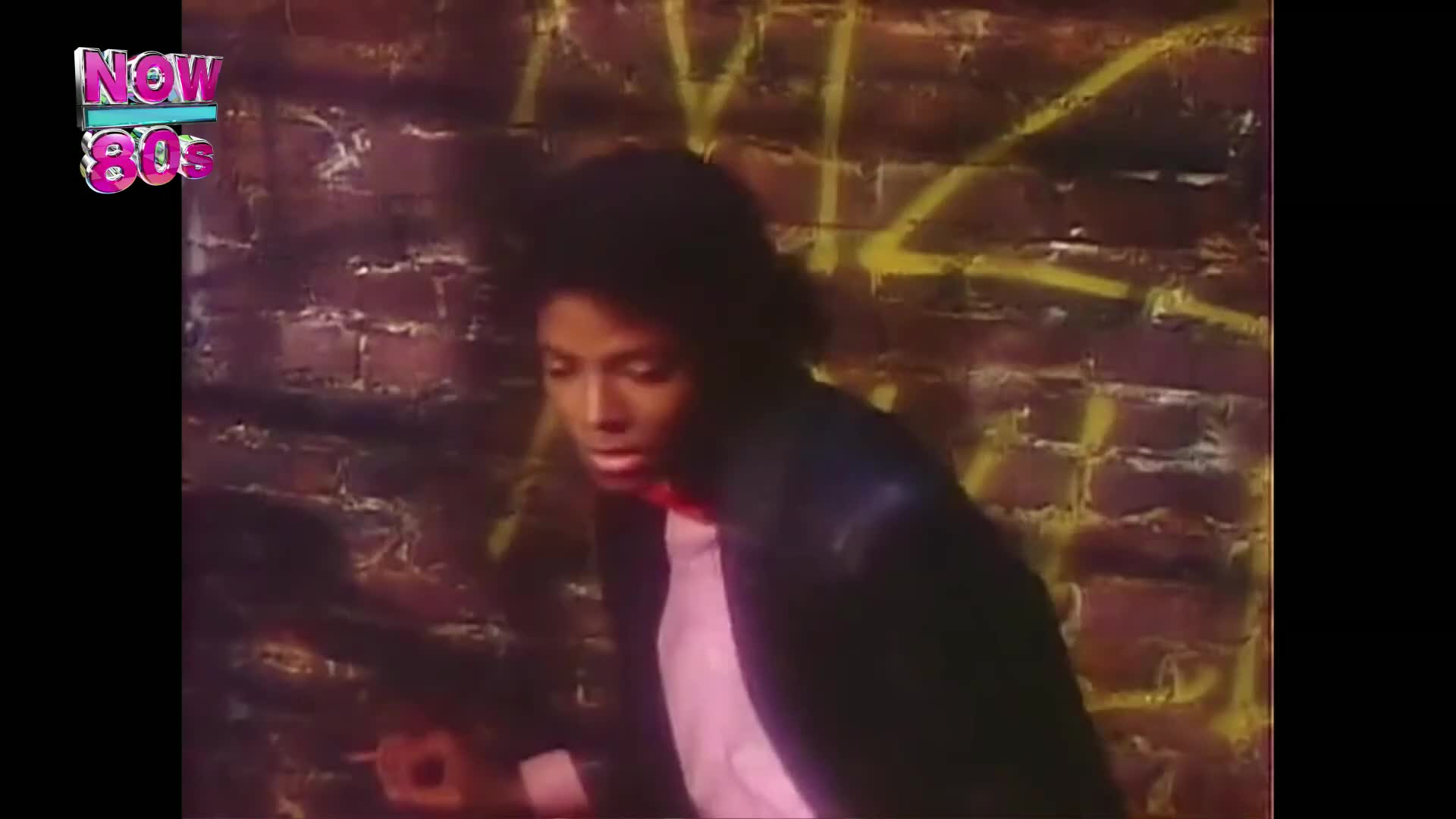 He's moving quickly, his dark hair flying as he turns toward the camera. Behind him, a brick wall is marked with yellow graffiti.
