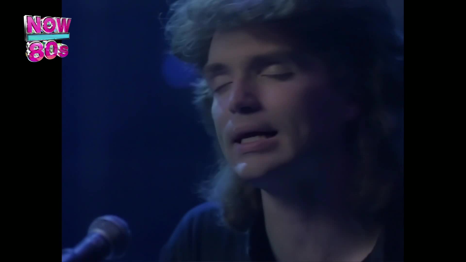 The singer, bathed in blue light, closes his eyes and sings into a microphone. His hair, a mass of blonde curls, frames his face as he performs.
