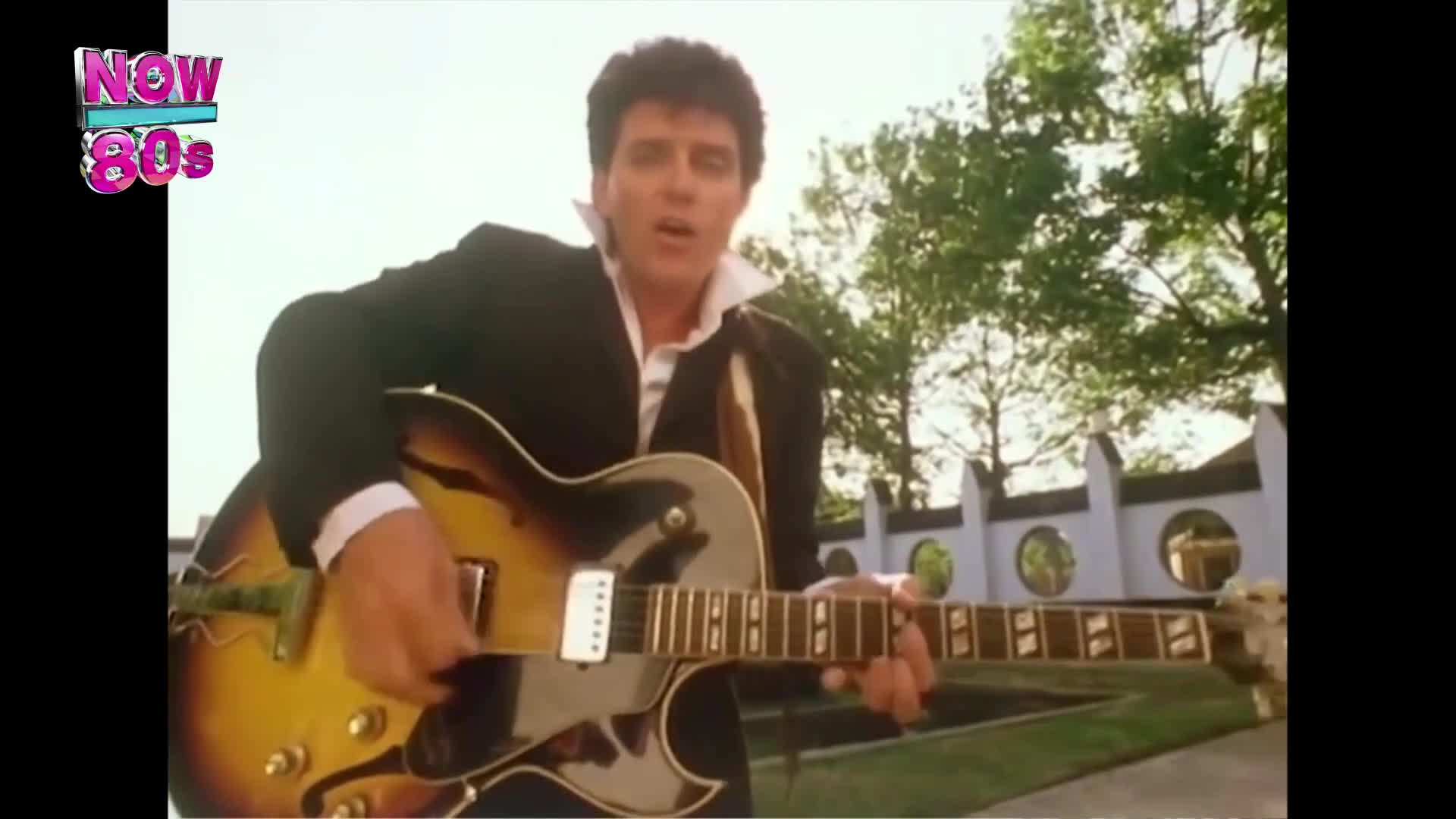 A man in a suit strums a sunburst guitar, singing directly into the camera. Behind him, a white picket fence and some trees are visible in the bright sunlight.

