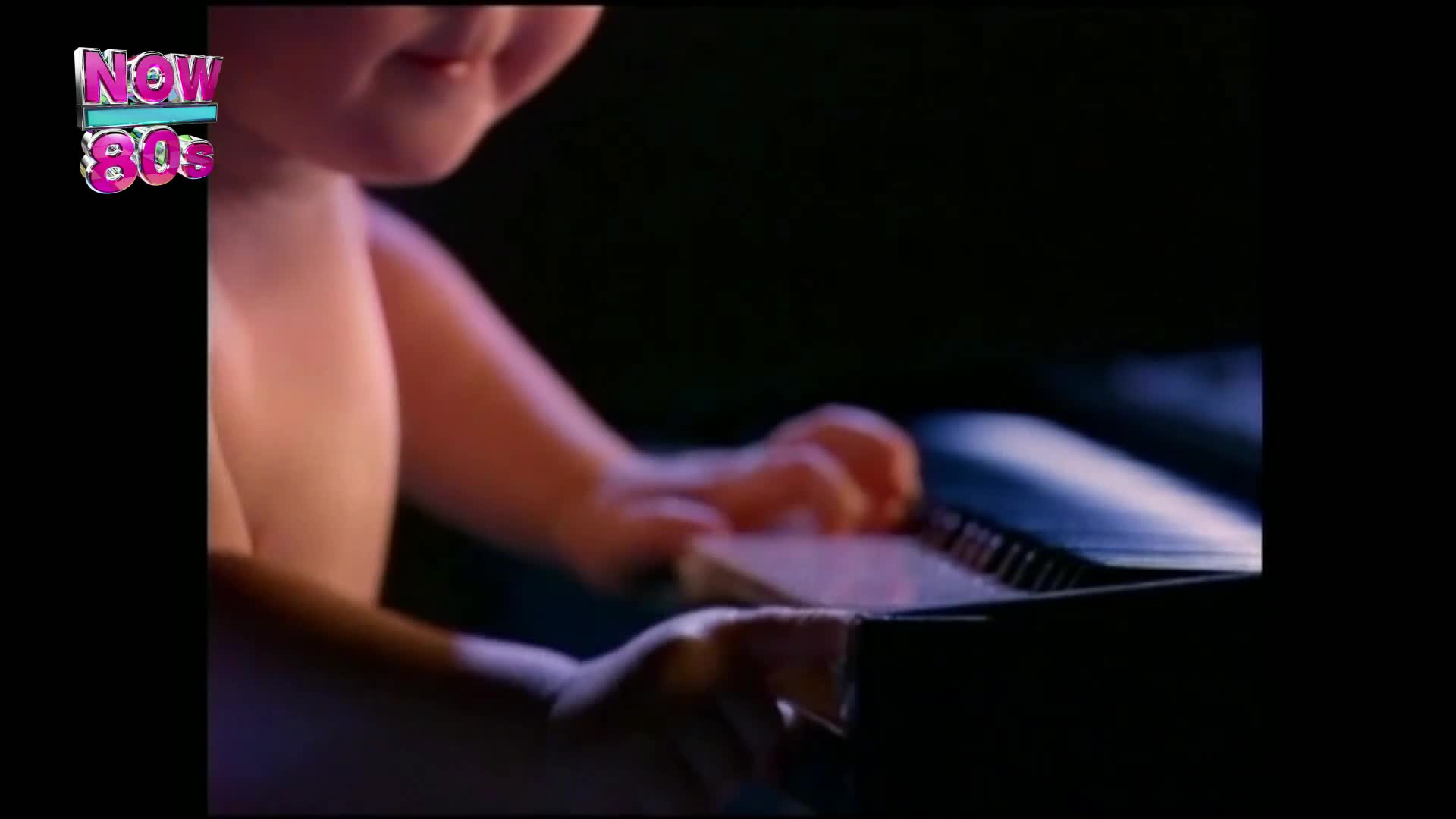 A child's hands press down on the keys of a synthesiser, the pinkish glow of the "Now 80s" logo in the corner hinting at the era. The dark background swallows the rest of the scene, focusing all attention on the small fingers making music.