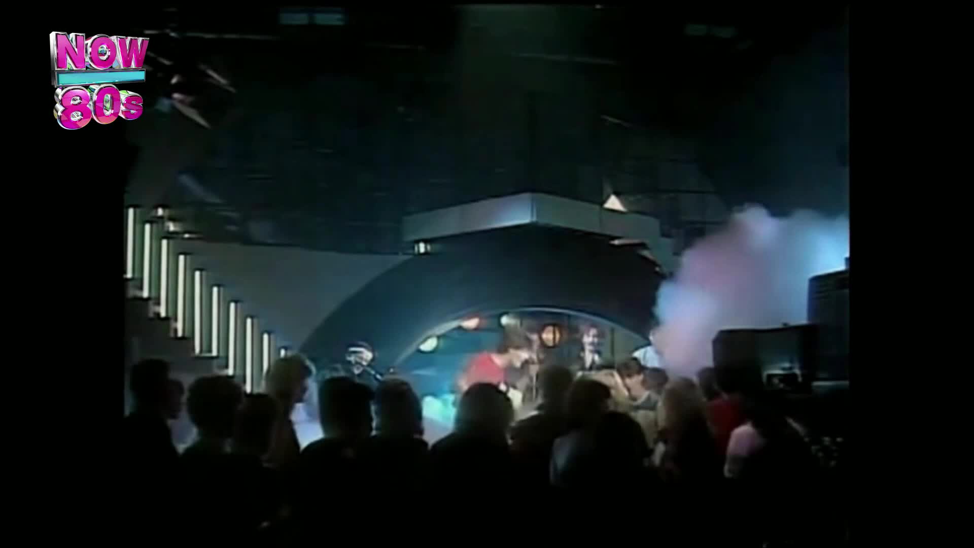 A band plays on a stage, bathed in dramatic lighting. Smoke billows around them as the crowd watches intently.