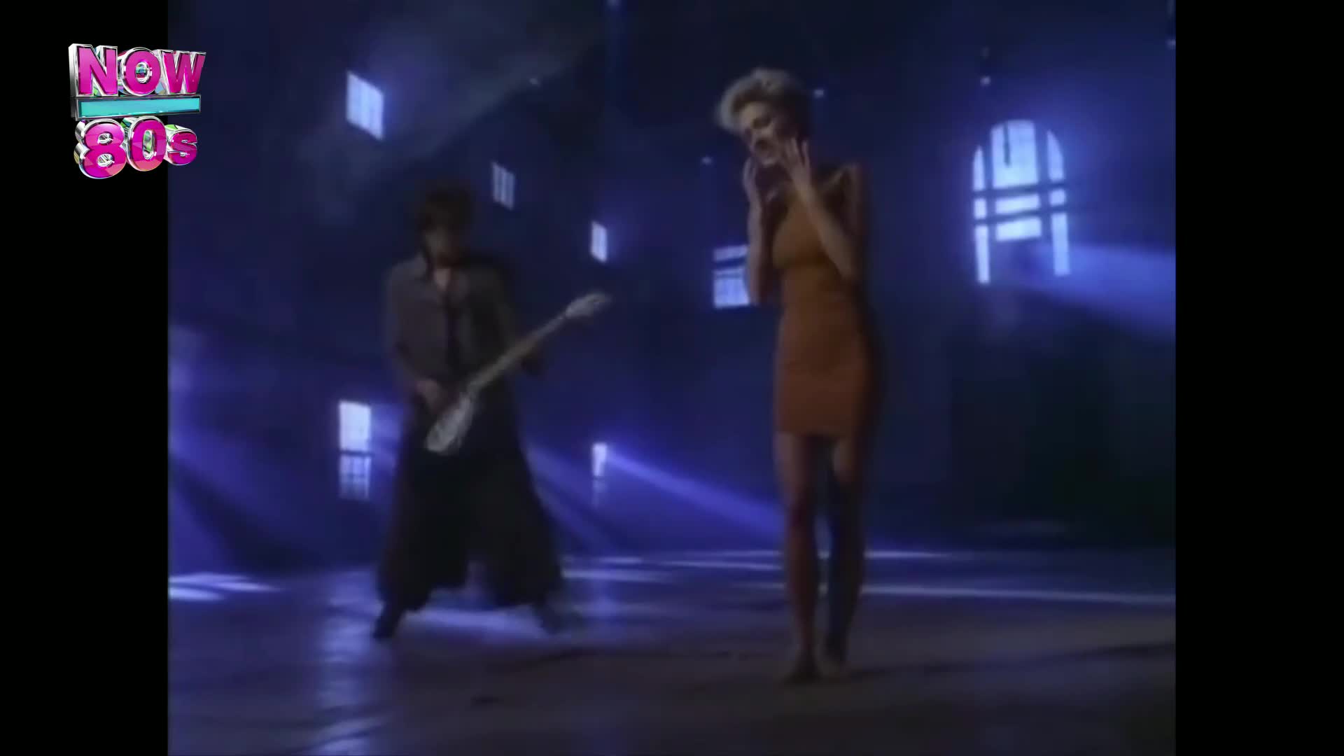 A man in dark, wide-legged trousers strums a white electric guitar, his body angled towards the camera. Beside him, a woman in a short, rust-coloured dress stands with her hands near her face, her blonde hair catching the blue stage lights.