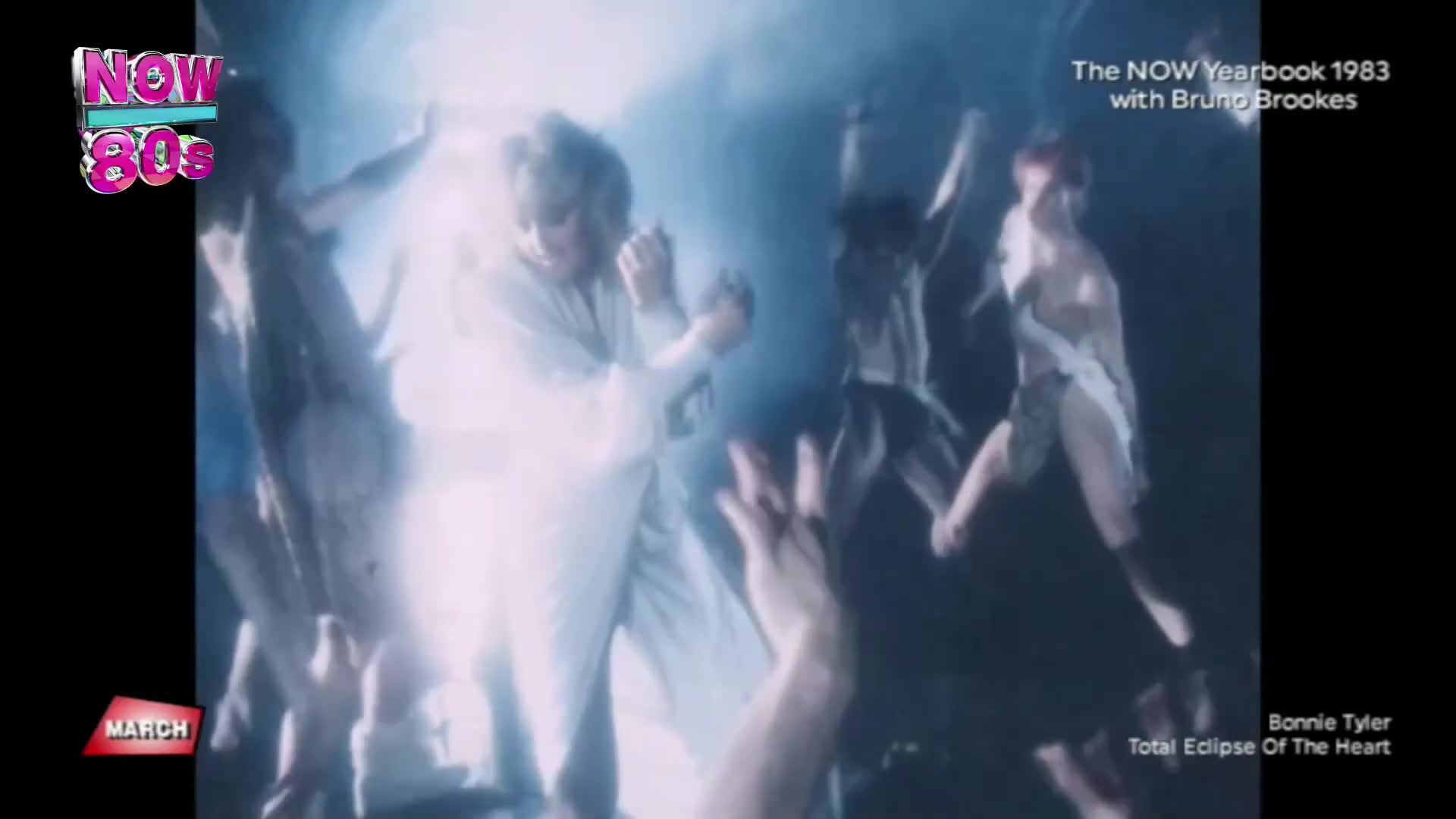 A group of dancers in white and tribal-inspired outfits move energetically under bright, hazy lights. Their arms are raised, and their bodies twist and turn as they perform.