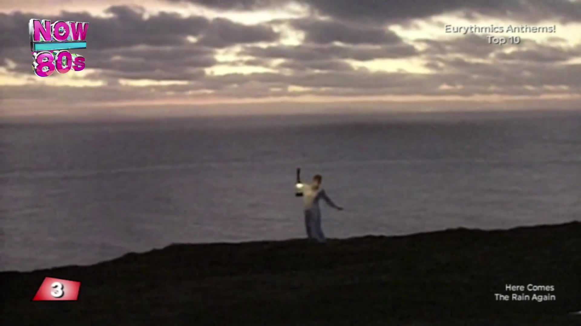 A lone figure stands on a cliff edge, holding a lantern aloft against a dramatic sky. The vast expanse of the sea stretches out before them, a muted grey under the fading light.