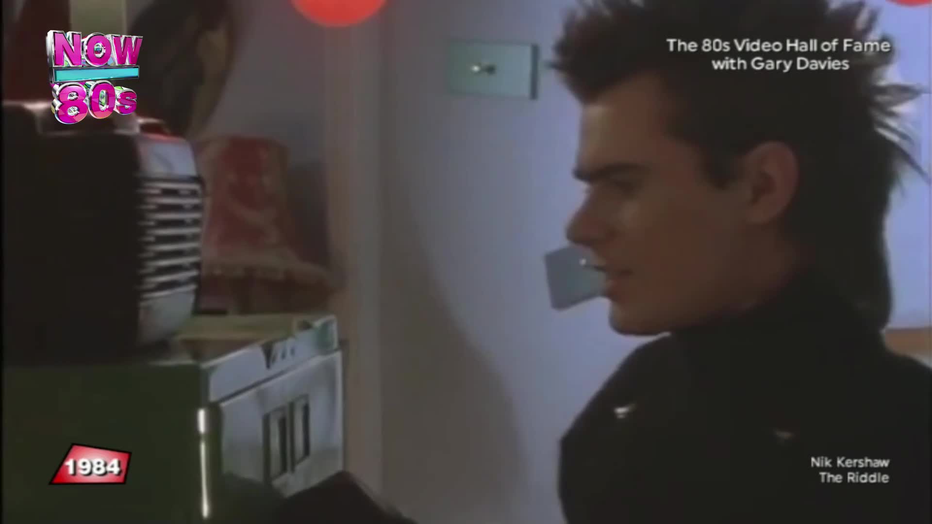 A young man with a spiky haircut turns his head, his gaze fixed on something off-camera. Beside him, a vintage cassette player sits on a green box, the year 1984 emblazoned on a red marker.