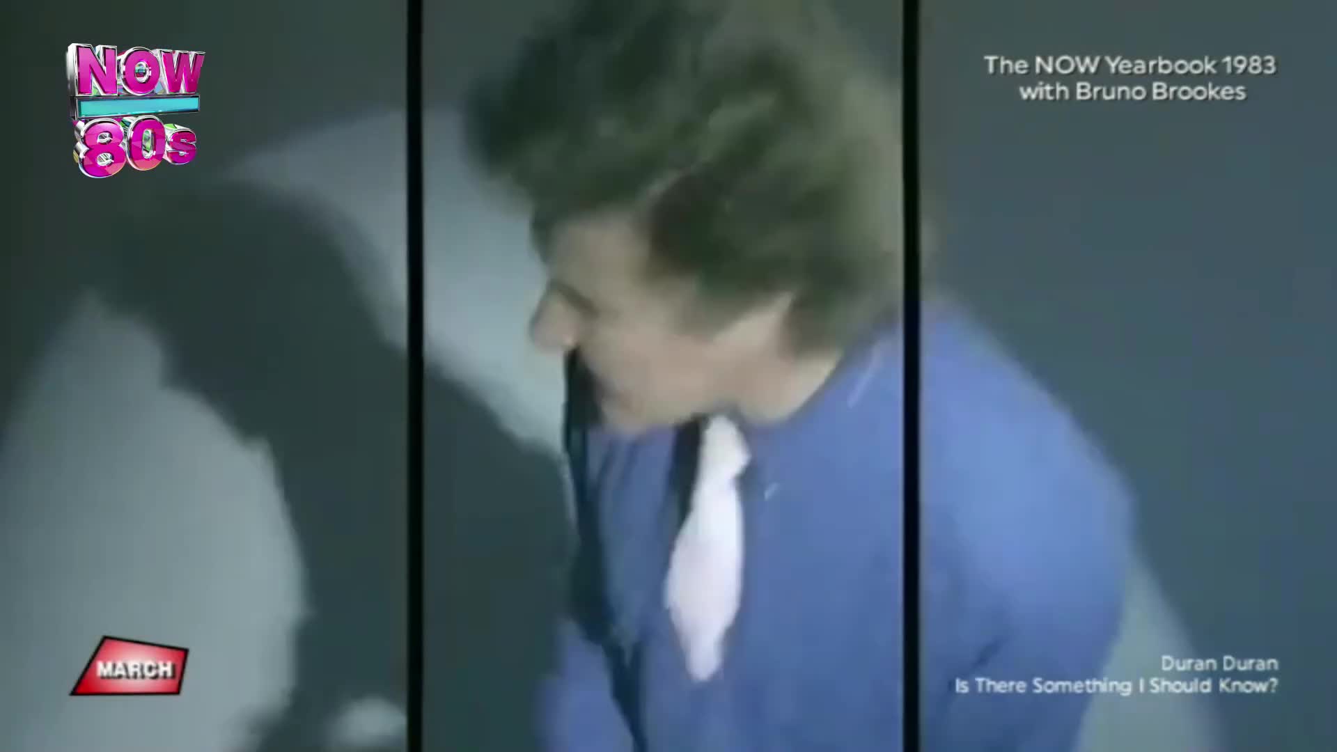 A man with big, styled hair in a blue jumper and white tie leans forward. The "NOW 80s" logo flashes in the corner.