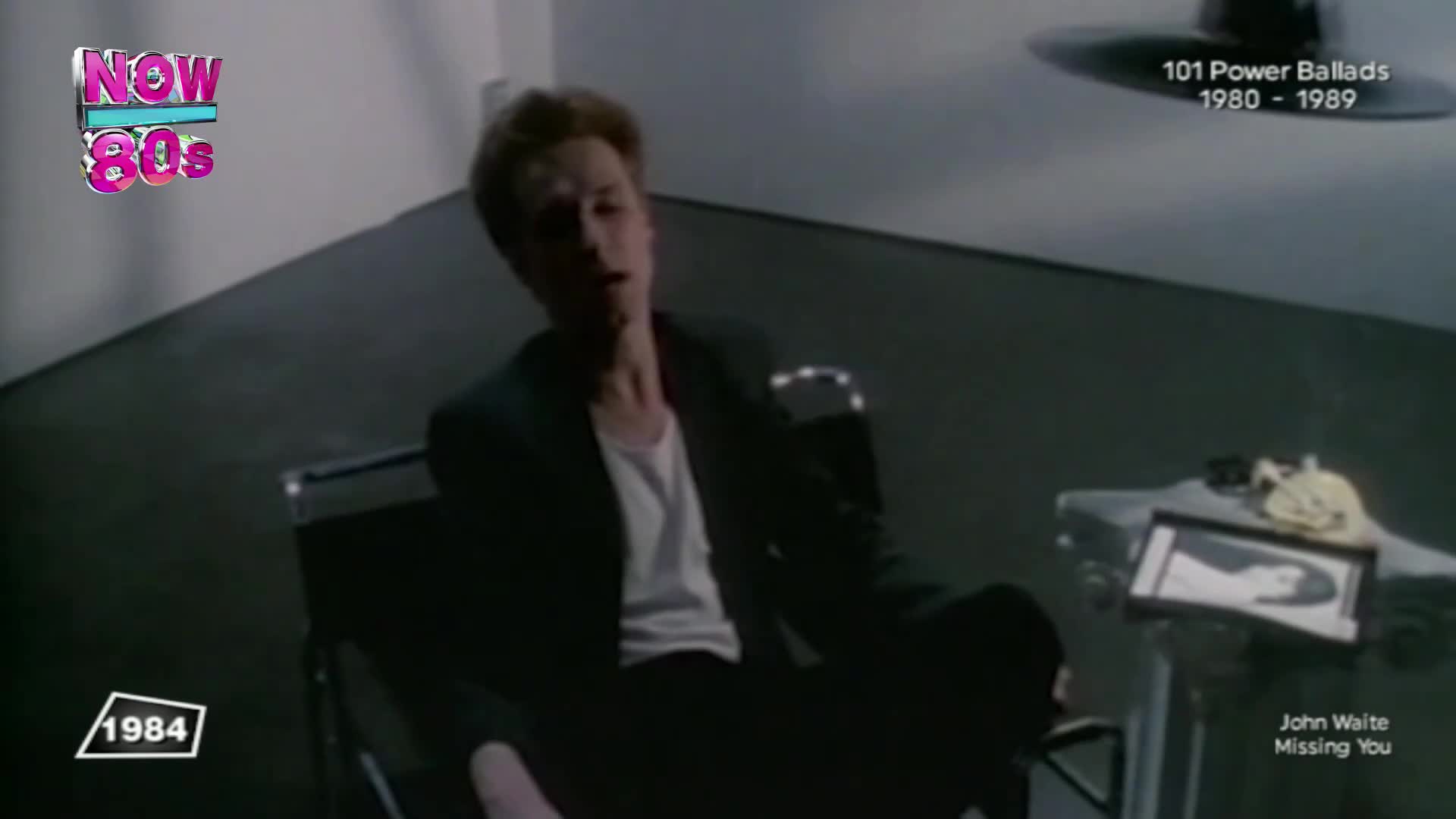 A young man in a dark suit slumps in a chair, his head tilted back. A framed photograph sits on a nearby table, hinting at the song's theme.