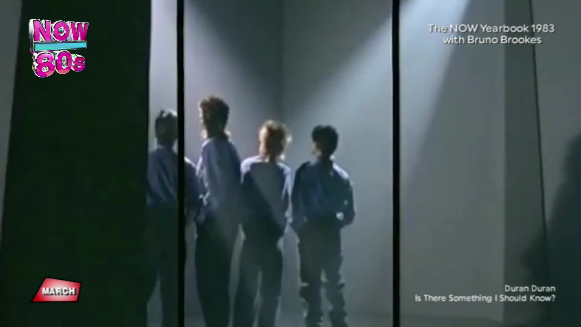 Four lads in matching blue shirts stand in a stark, spotlighted room. They're looking off to the side, their faces obscured by shadow and the grainy quality of the recording.