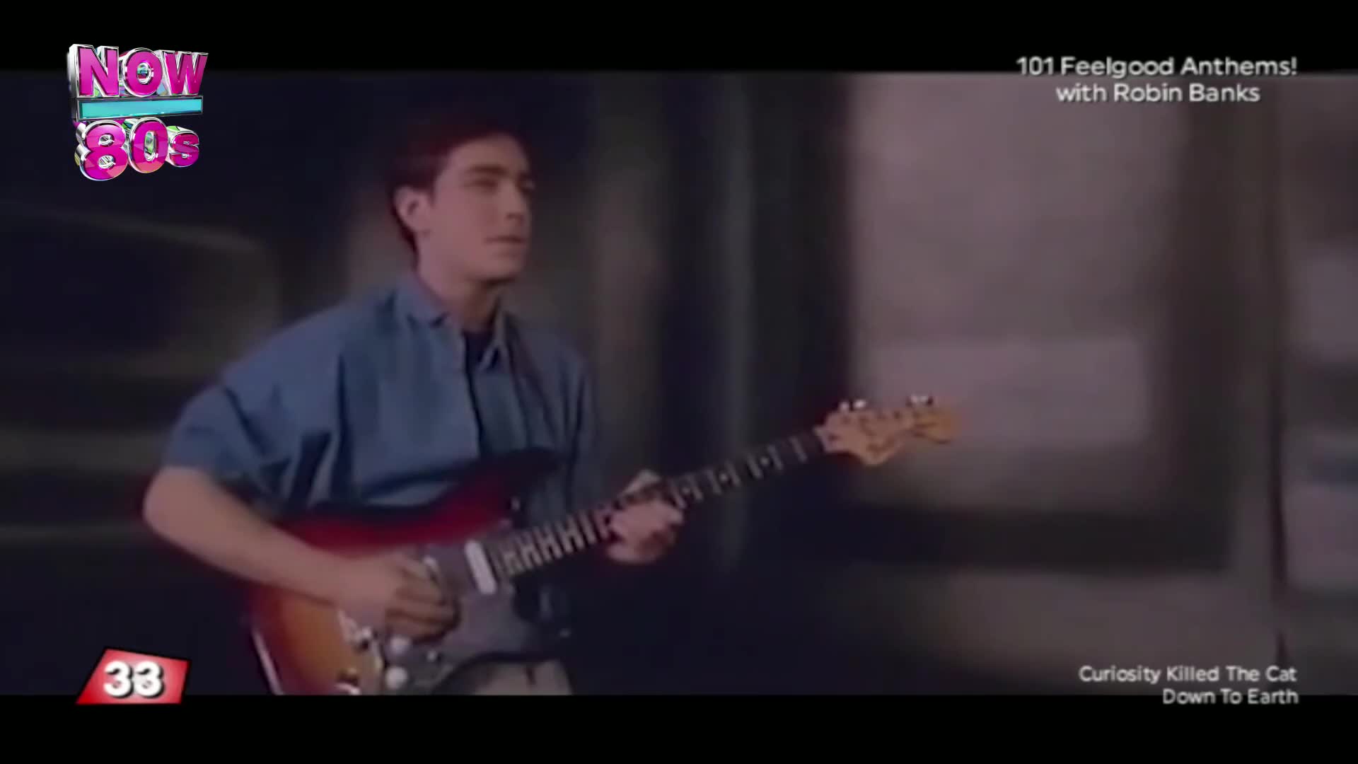 A young man in a blue shirt plays a sunburst electric guitar. The music video is from the 80s, featuring the band Curiosity Killed the Cat.