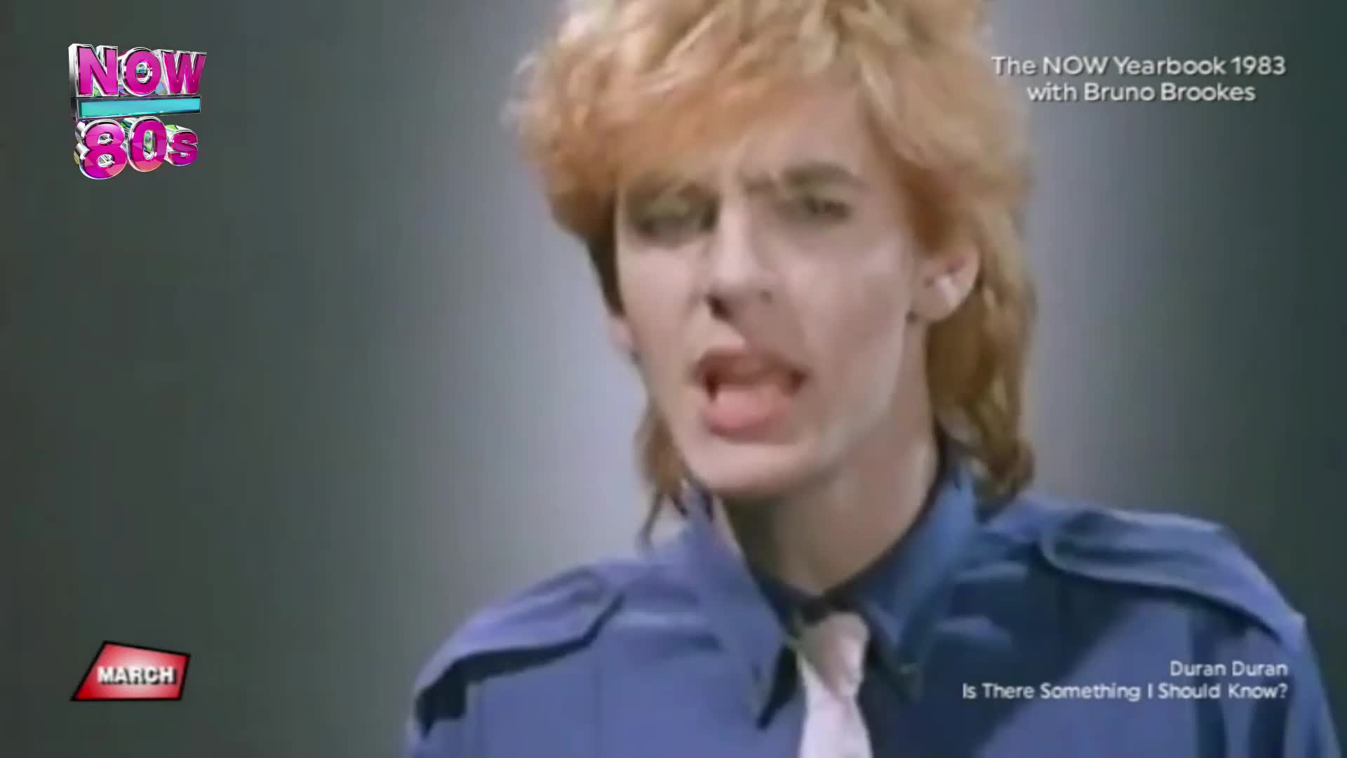 A man with feathered blonde hair, wearing a blue shirt and tie, sings into a microphone. The NOW 80s logo and "The NOW Yearbook 1983 with Bruno Brookes" text appear on screen.