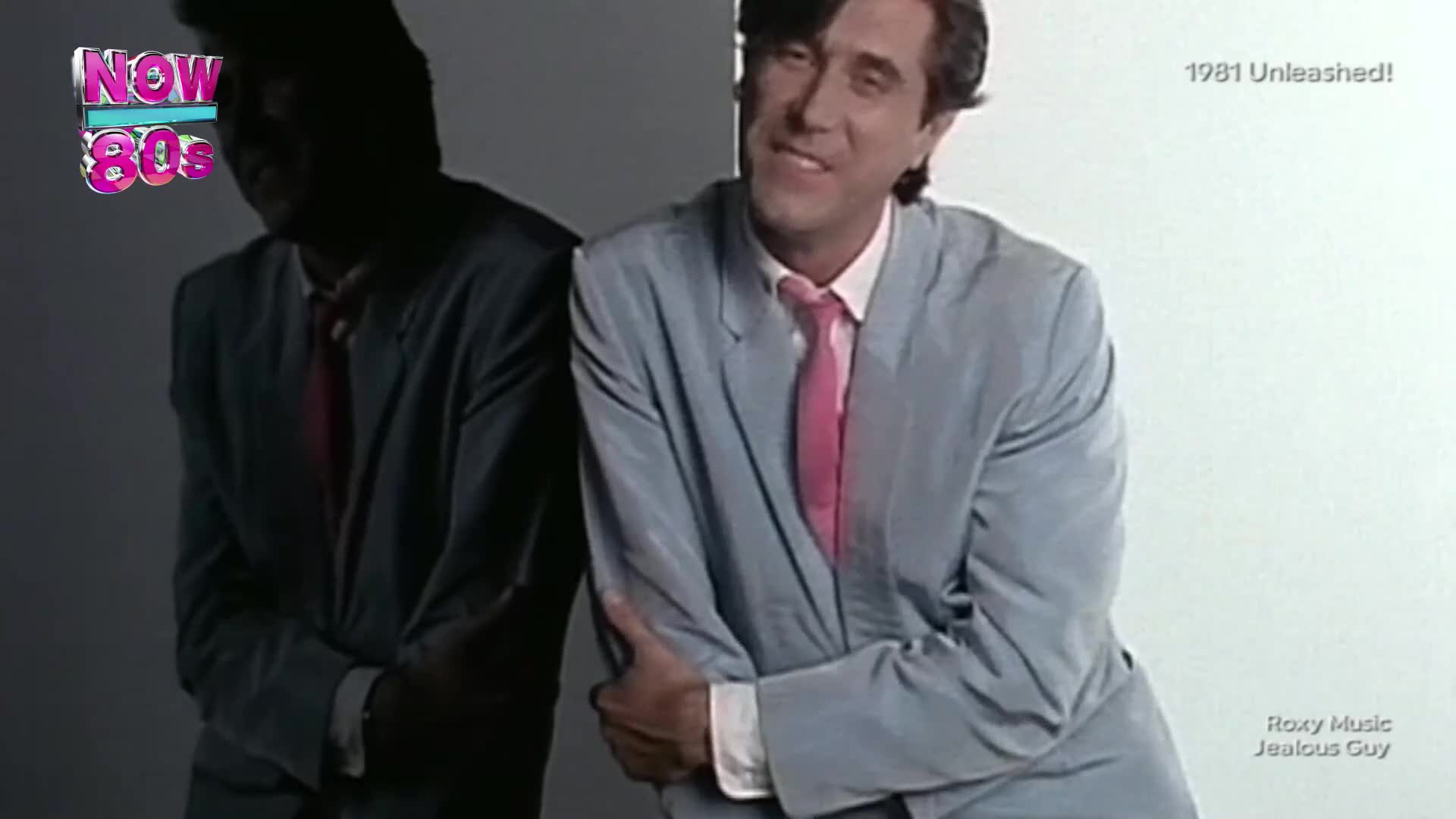 A man in a light blue suit and pink tie leans against a reflective surface, his expression a wry smile. The "NOW 80s" logo hovers in the background, hinting at a classic British music video from 1981.