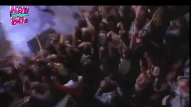 A huge crowd surges forward, hands raised in unison.  The air is thick with smoke, and the "Now 80s" logo flashes in the corner.