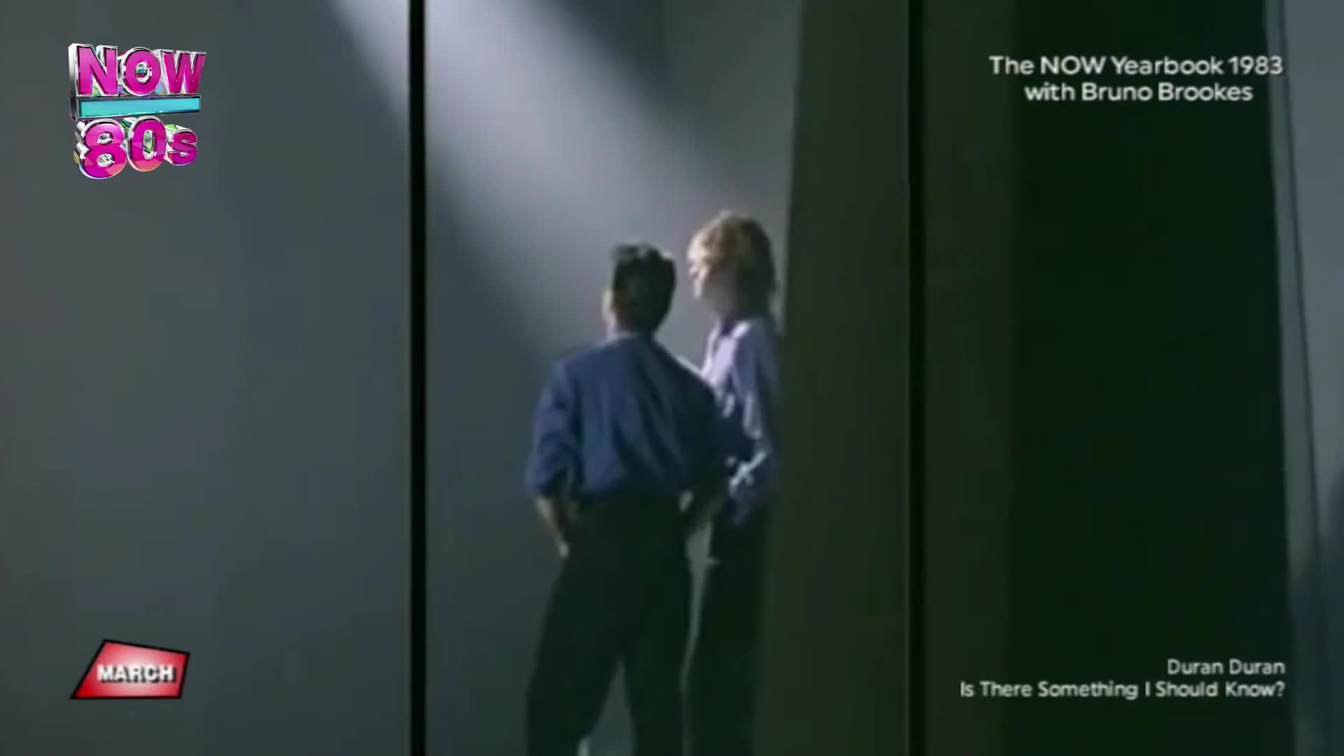Two men stand in a stark, dimly lit space, their backs to me. One wears a dark blue shirt and trousers, the other a lighter purple shirt and dark trousers. A bright spotlight hits them from above.