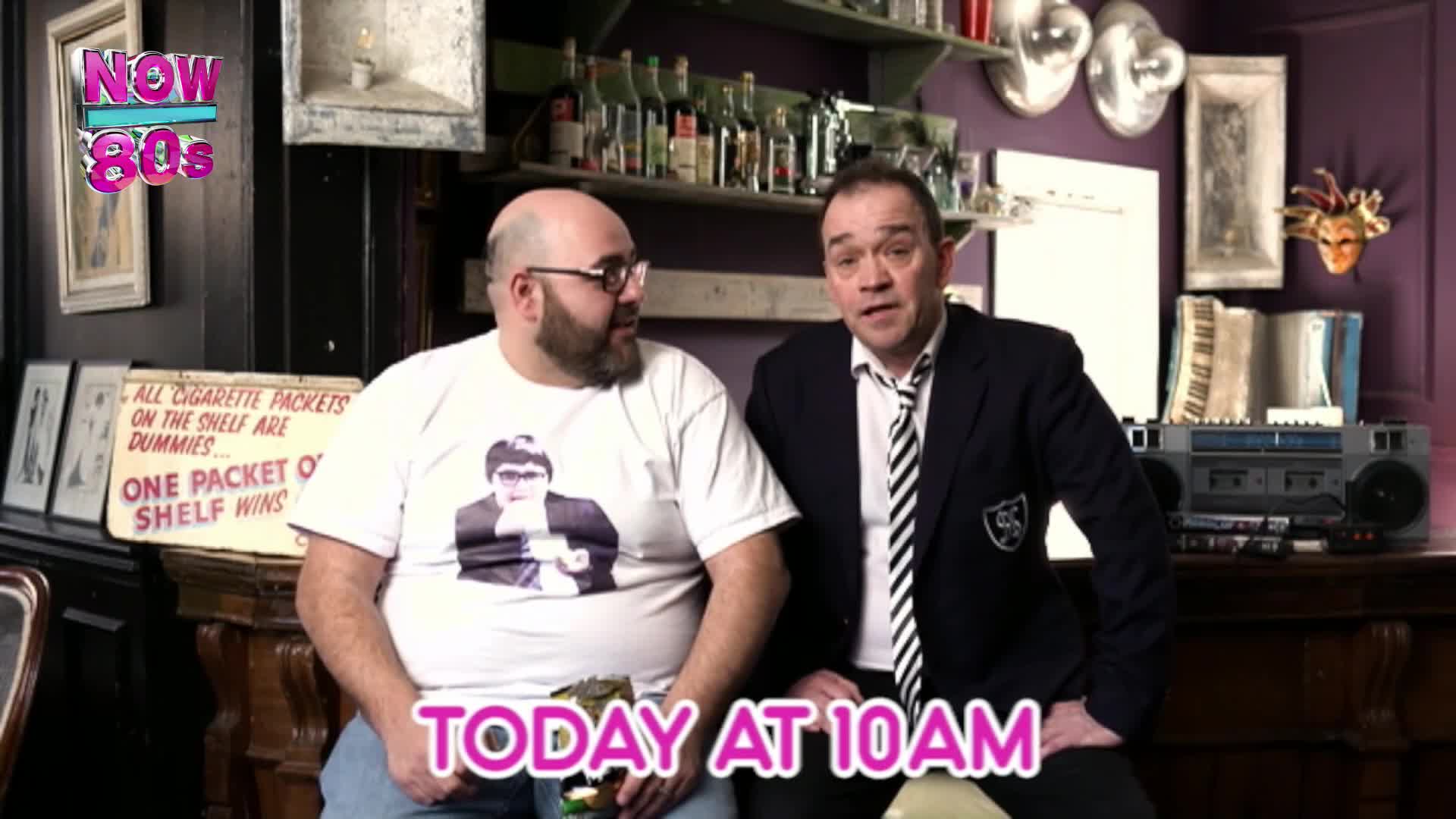 Two men are sitting in what looks like a pub, one in a t-shirt with a picture on it, the other in a blazer. A boombox sits on a shelf behind them, hinting at the era.