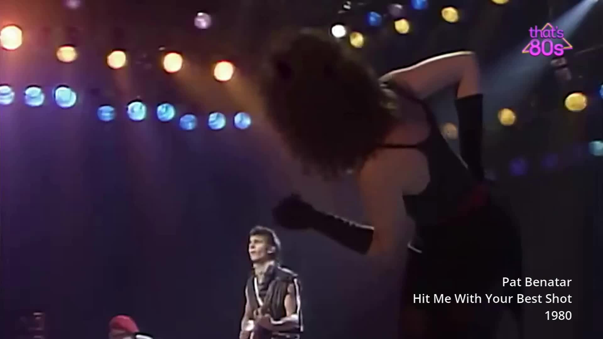 The lead singer, Pat Benatar, throws her head back as she sings, her dark hair flying. A guitarist in a sleeveless vest stands to her left, bathed in the stage lights. The lead singer, Pat Benatar, throws her head back as she sings, her dark hair flying. A guitarist in a sleeveless vest stands to her left, bathed in the stage lights.