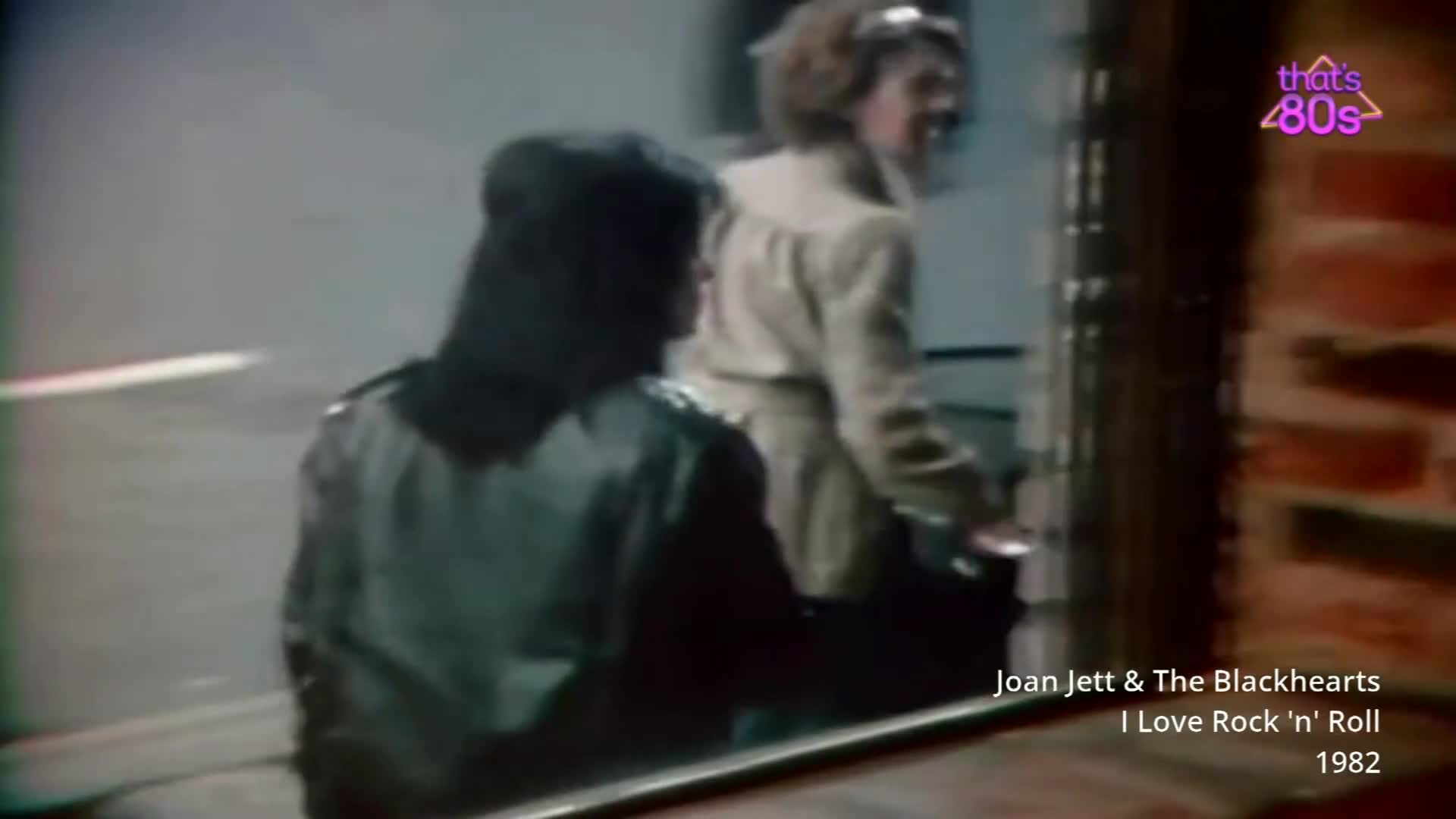 A woman in a dark jacket turns her head, her black hair falling forward. Another woman in a light-coloured coat walks past a brick wall. A woman in a dark jacket turns her head, her black hair falling forward. Another woman in a light-coloured coat walks past a brick wall.